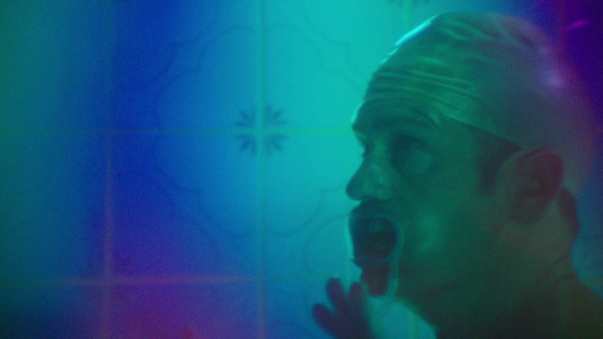 Mann in einer Dusche mit Badekappe und offenem Mund. Blaue und türkisfarbene Fliesenwand im Hintergrund.
