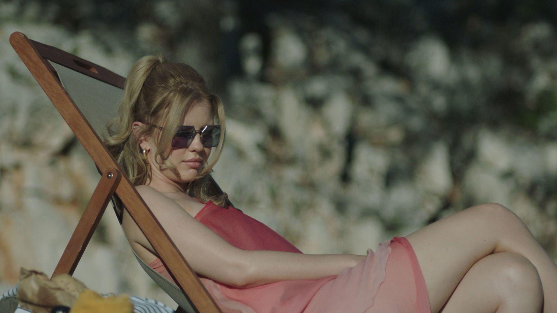 Frau mit Sonnenbrille, rotem Kleid, liegt auf einem Strandstuhl. Sonnenlicht und Bäume im Hintergrund, entspannte Szene.