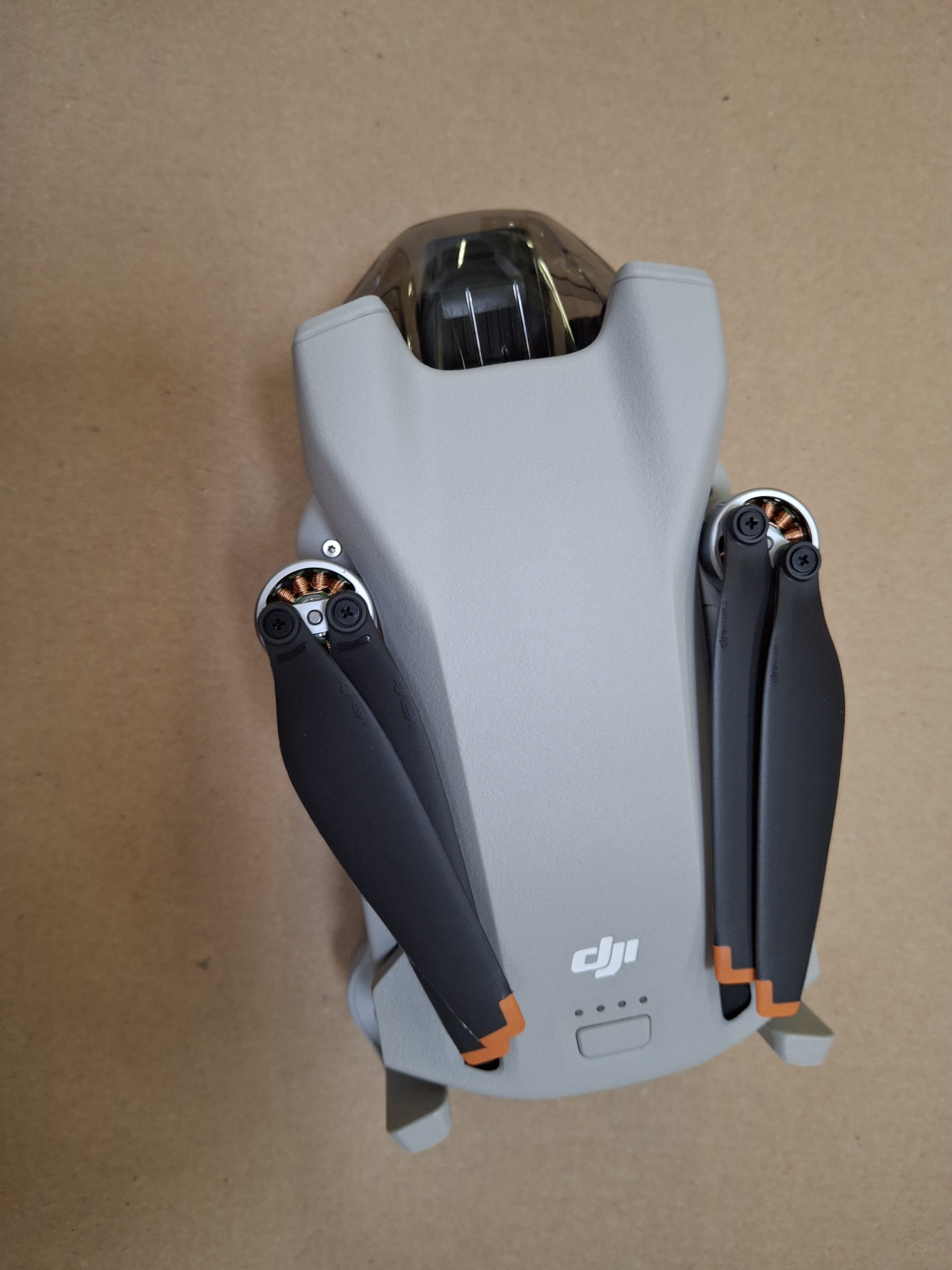 Szary dron ze złożonymi ramionami, czarnymi śmigłami i pomarańczowymi końcówkami. Leży na brązowej powierzchni.