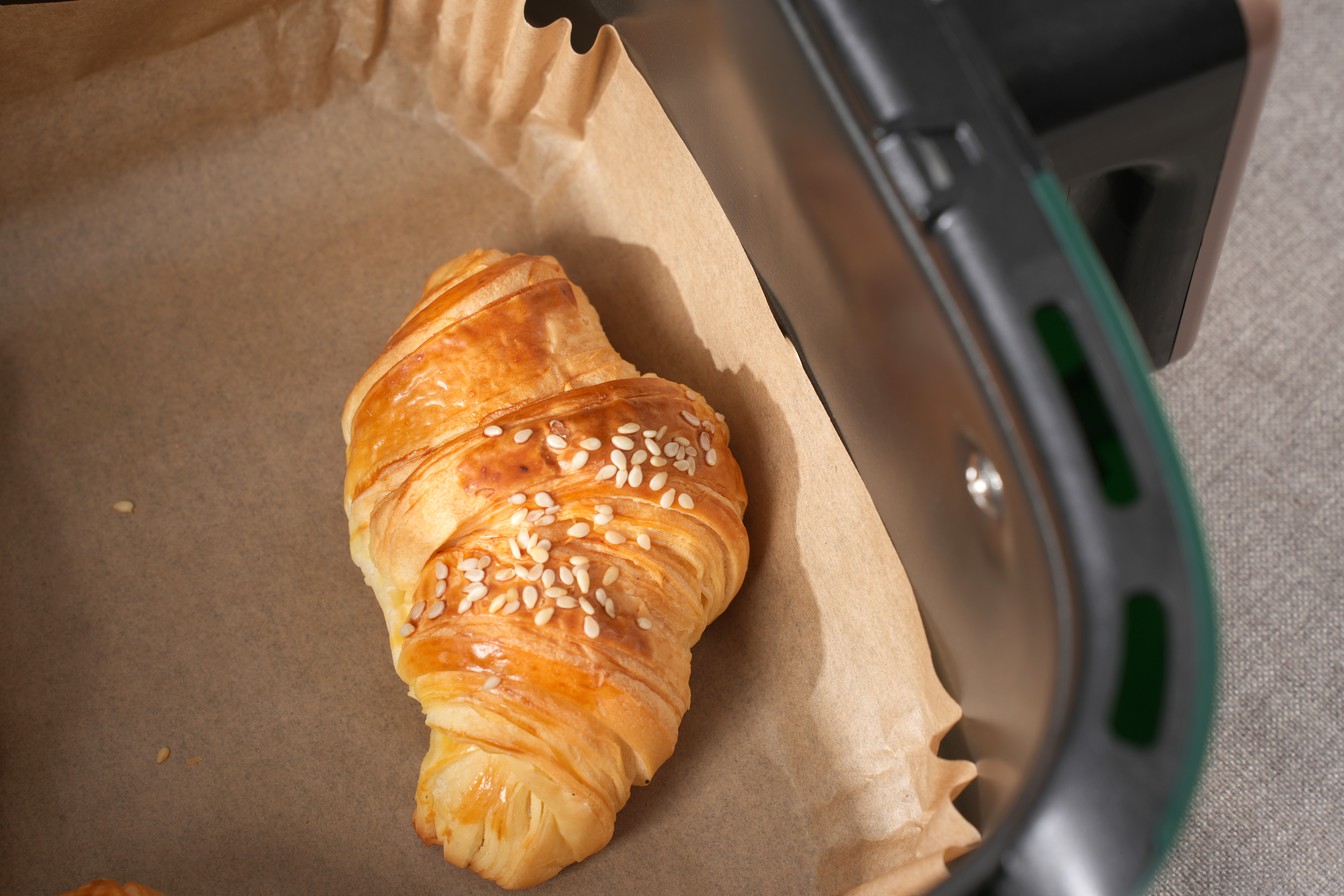 Croissant w blaszce do pieczenia. Brązowy, z nasionami sezamu. Taca jest w czarnej frytkownicy beztłuszczowej.