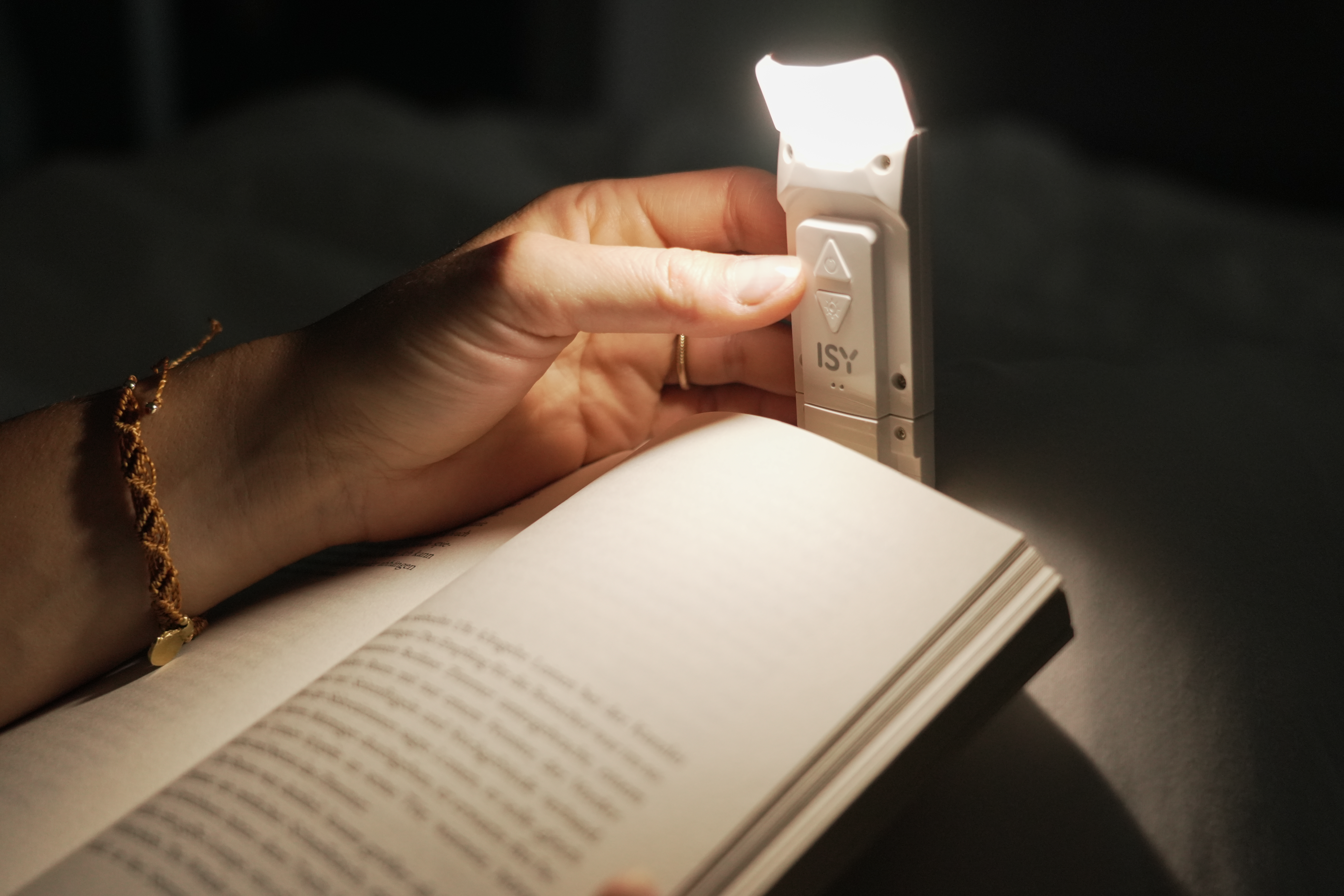 Une main tient une lampe de lecture au-dessus d'un livre ouvert. Un bracelet doré est visible.