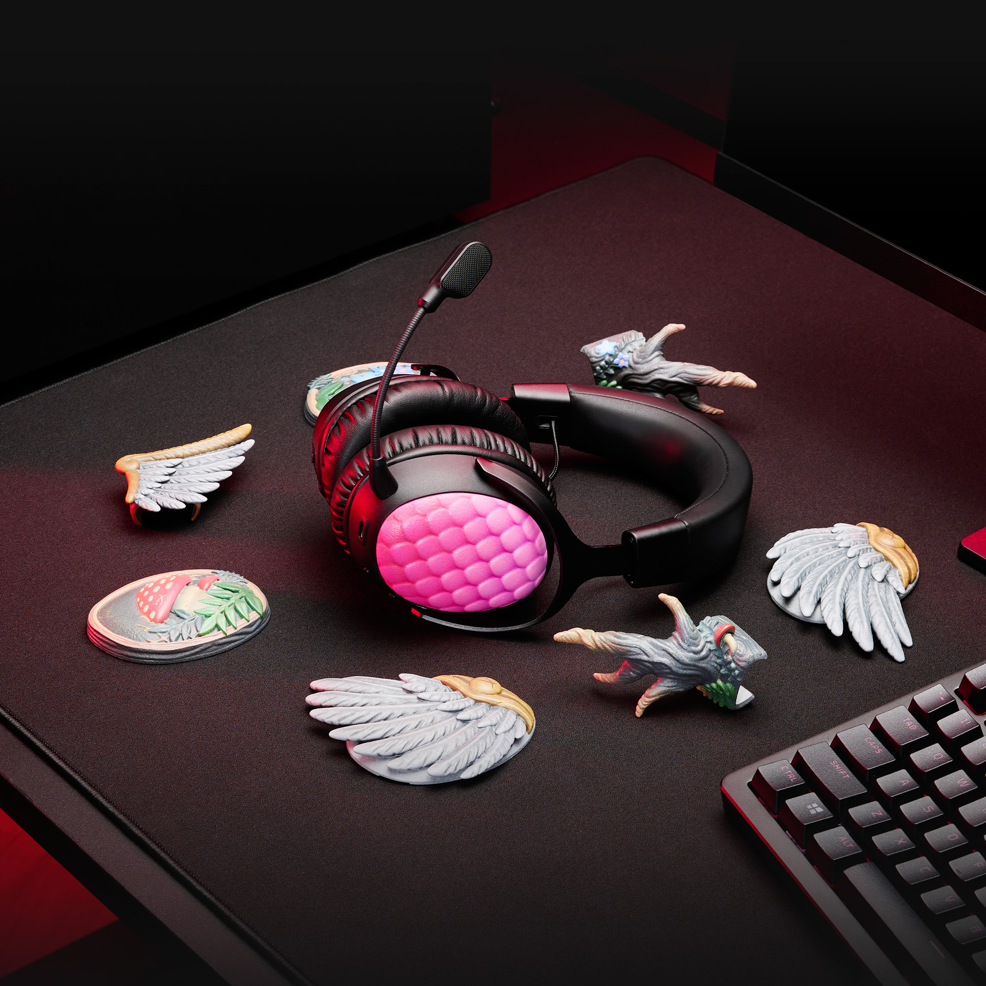 Schwarzes Headset mit rosa Ohrmuscheln, Engelsflügeln und Tastatur auf dunkler Oberfläche. Rote Beleuchtung akzentuiert die Szene.