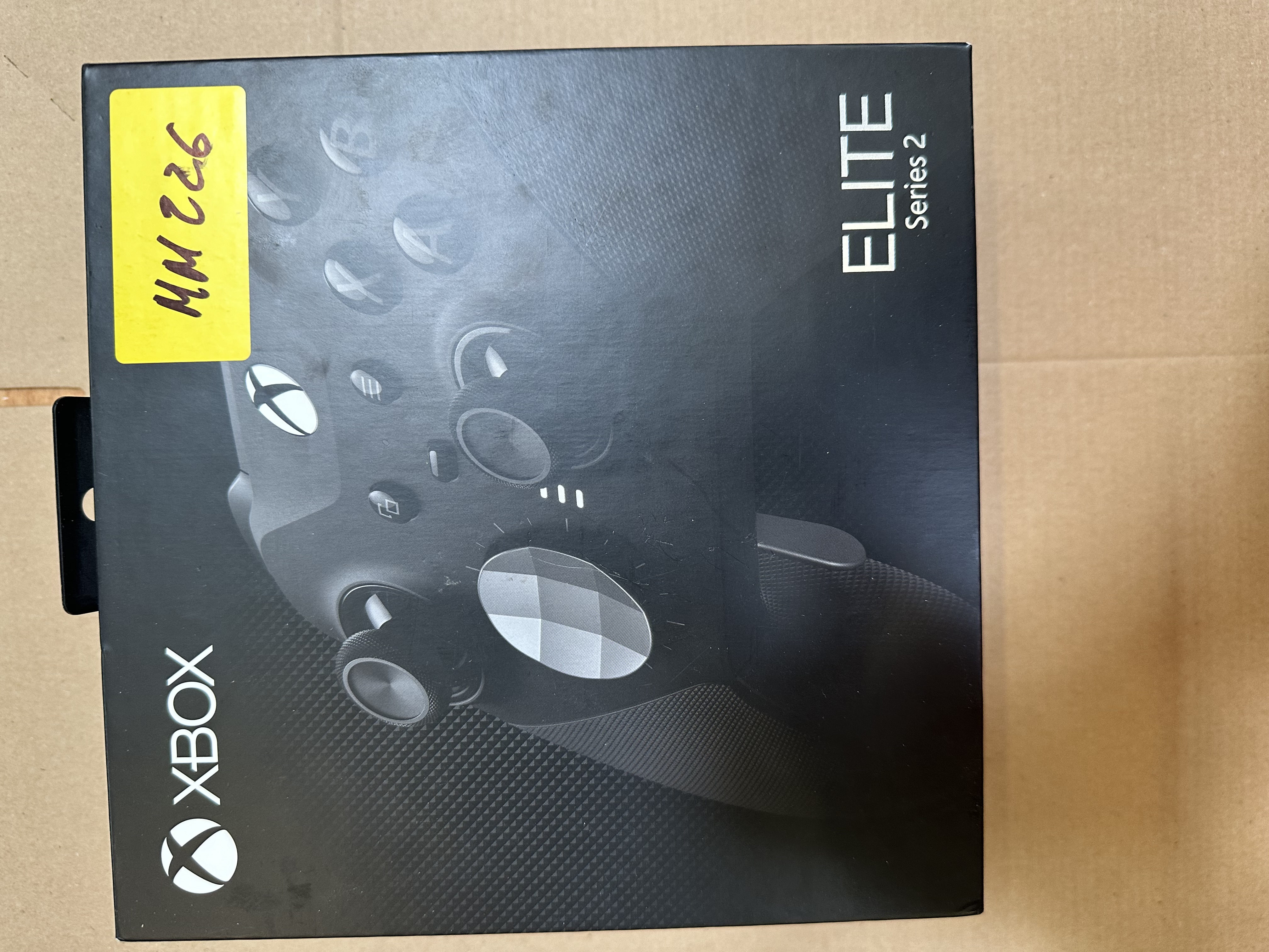 Czarne pudełko kontrolera Xbox Elite Series 2 z żółtą karteczką. Pudełko leży na kartonowej powierzchni.