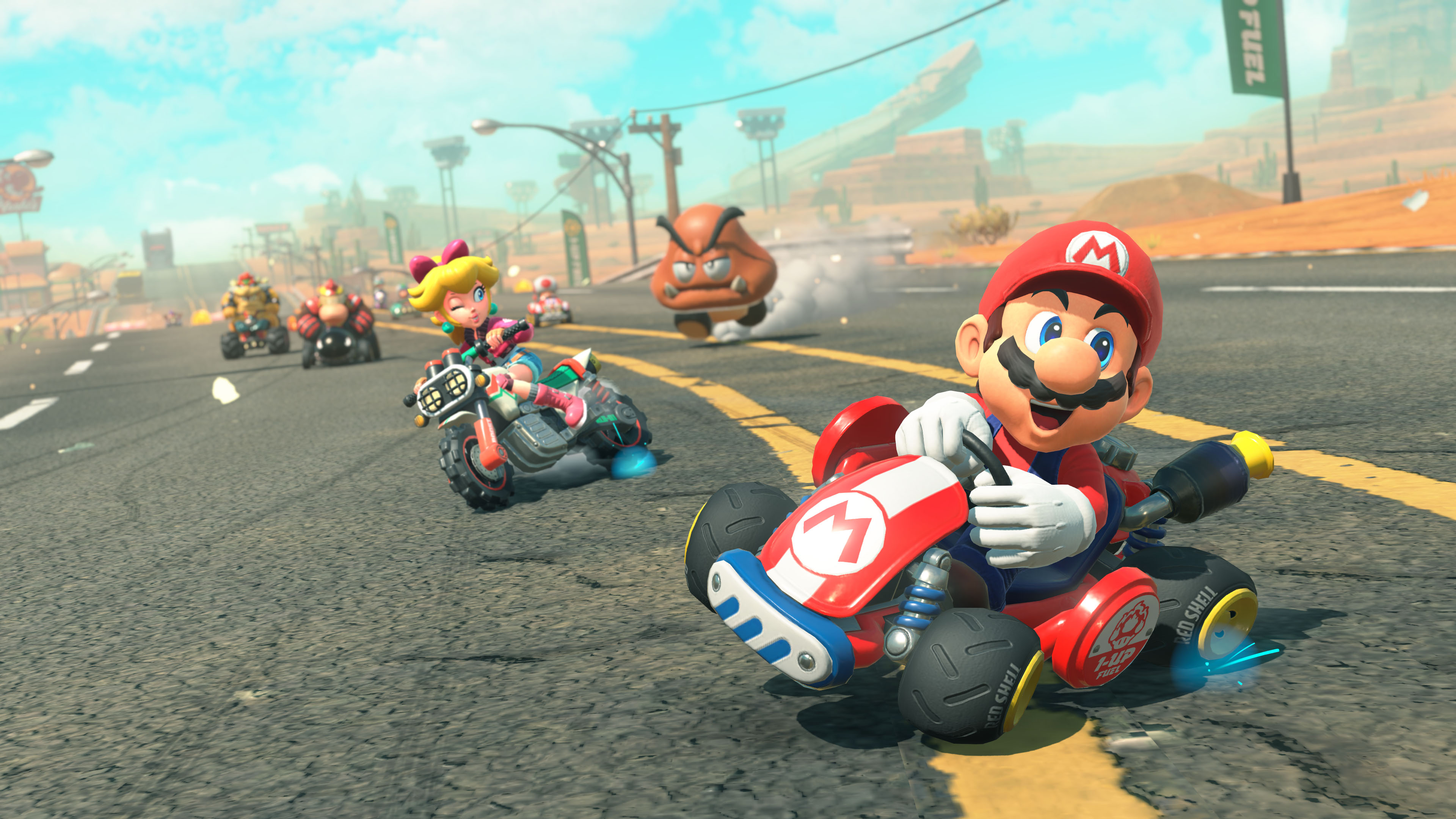 Mario Kart karakterleri, güneşli bir arka planla bir parkurda yarışıyor.
