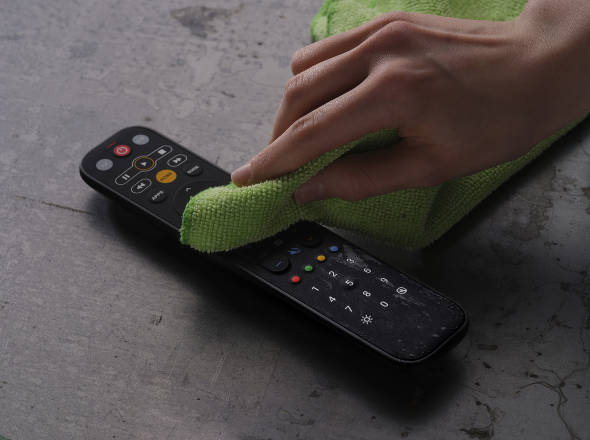Hand wischt schmutzige TV-Fernbedienung mit grünem Tuch ab. Fernbedienung hat viele Knöpfe. Graue Oberfläche.