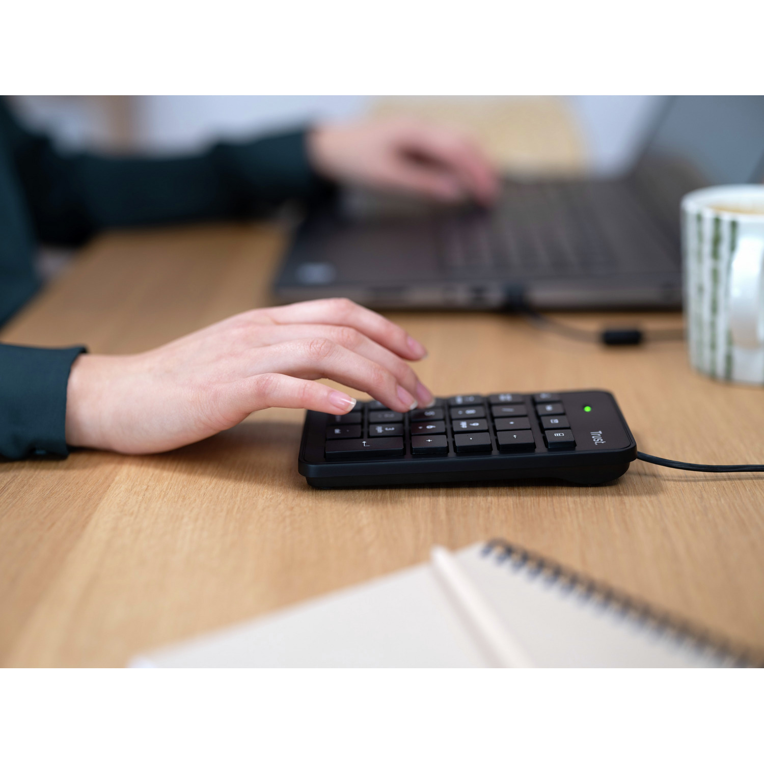 A person's hand using a numeric keypad on a wooden desk with a laptop and a mug.