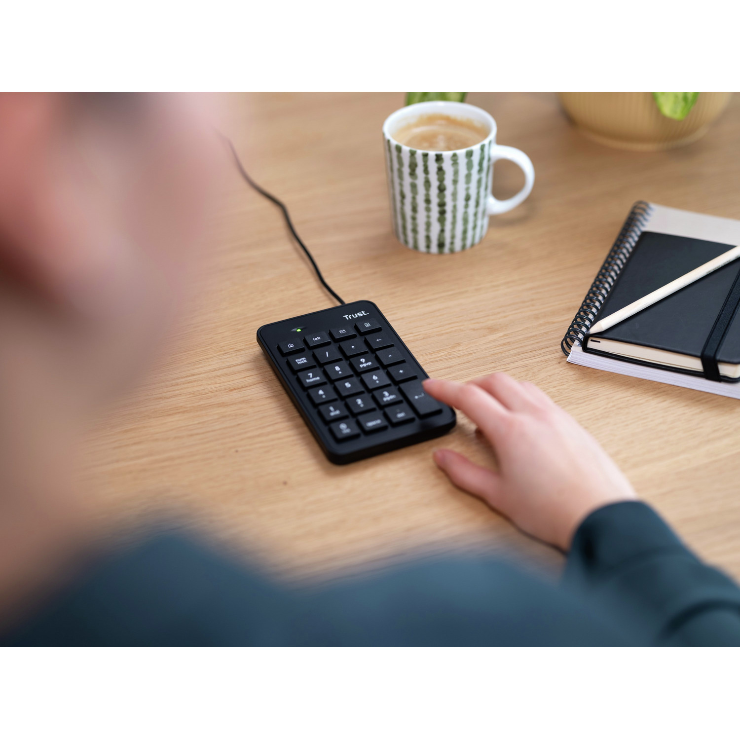 Person's hand on a black numeric keypad with a coffee mug and a notebook on a wooden desk.