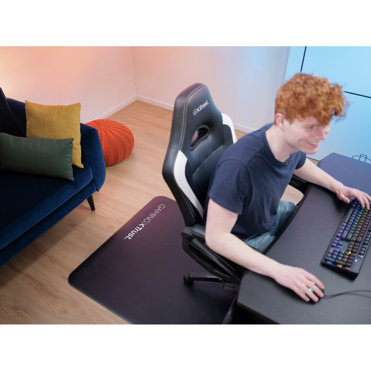 Man gaming with chair and mat. Black desk, blue sofa, orange ottoman, and wooden floor.