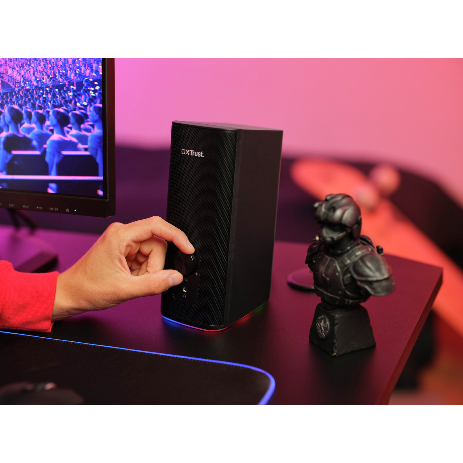 A person turns the volume knob on a black speaker. A bust is on the desk. Pink and red background.