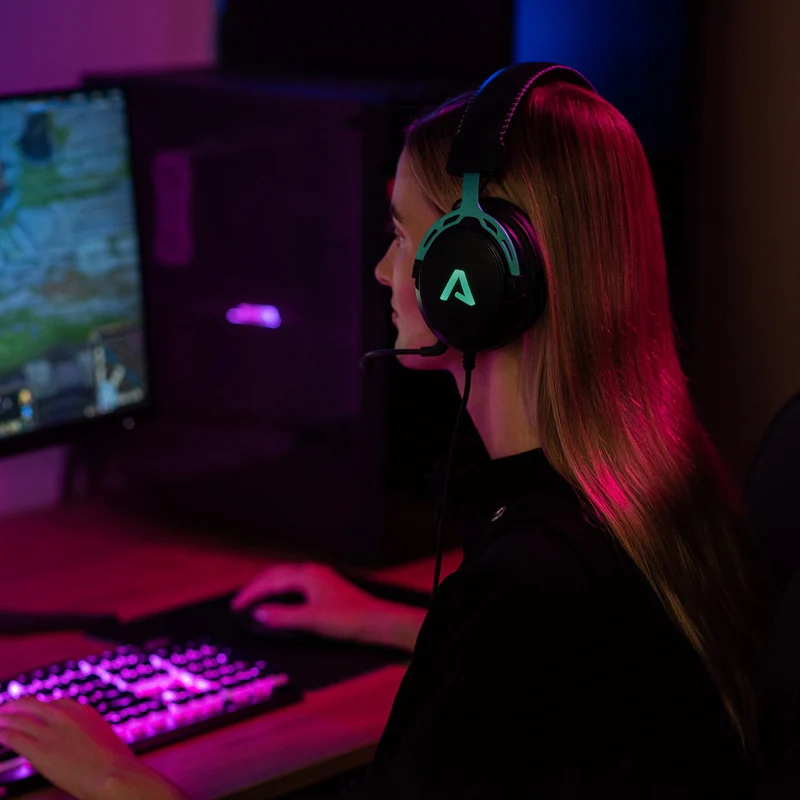 Nő videojátékot játszik fekete gamer headsettel, türkiz díszítéssel és világító logóval.