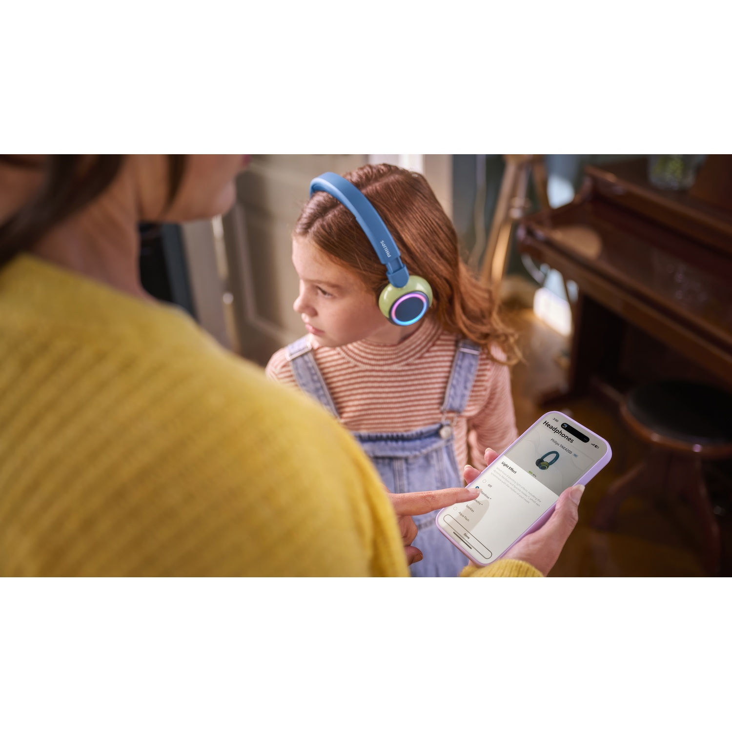 Una donna tiene in mano un telefono, mostrando a un bambino un paio di cuffie. Un pianoforte è sullo sfondo.