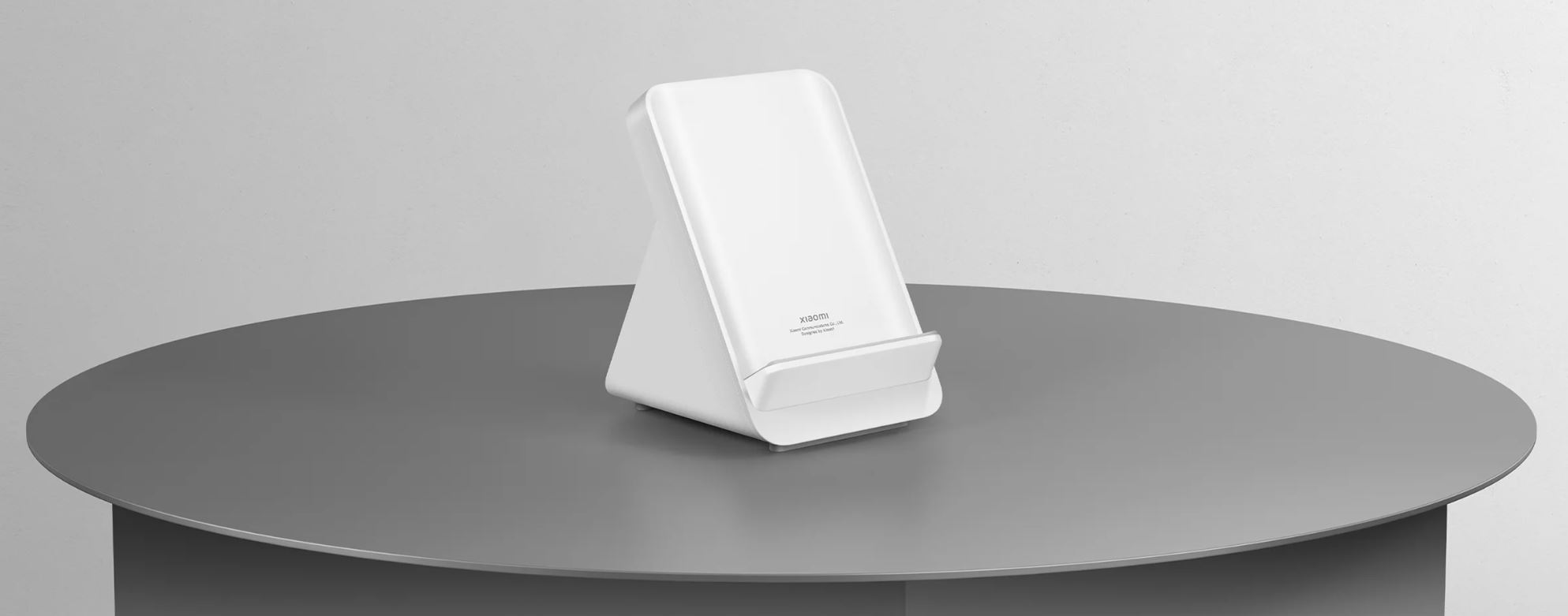 Biała ładowarka bezprzewodowa na szarym, okrągłym stole. Ładowarka stoi pionowo z logo Xiaomi.