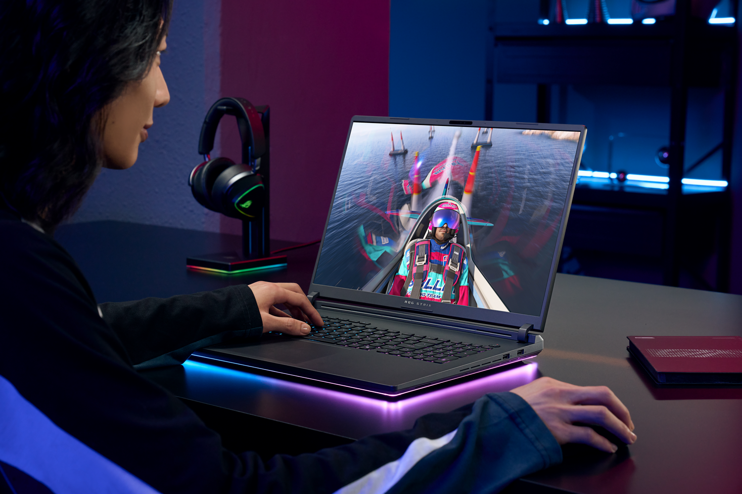 Una persona usa un portátil con un juego en la pantalla. Auriculares en un soporte a la izquierda.