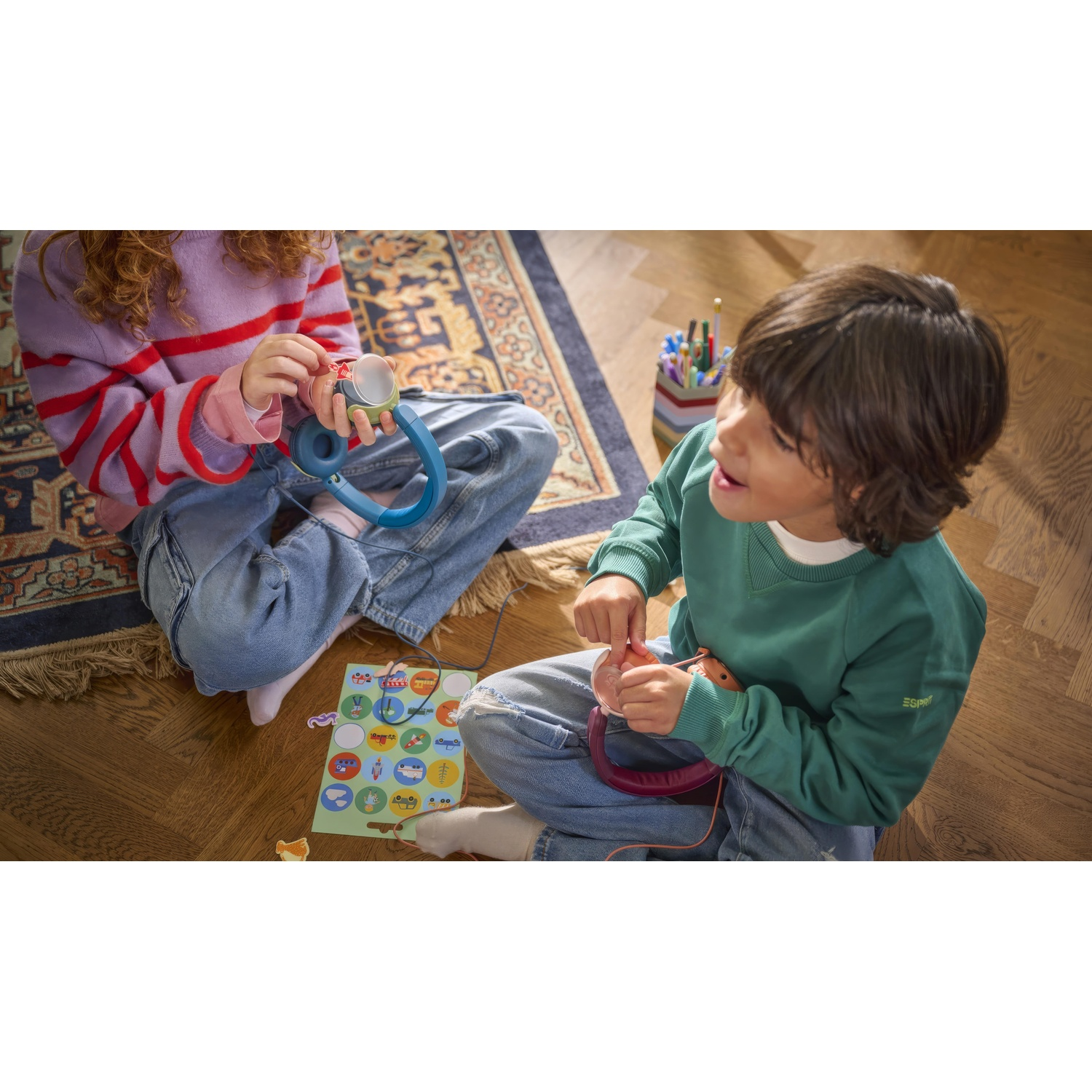 Due bambini seduti. Uno tiene cuffie blu, l'altro rosse. Adesivi colorati e pavimento in legno visibili.