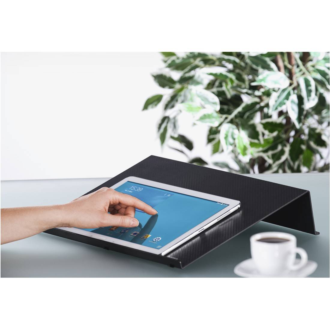 Hand berührt Tablet auf schwarzem, abgewinkeltem Ständer. Eine Tasse Kaffee steht auf dem Tisch.