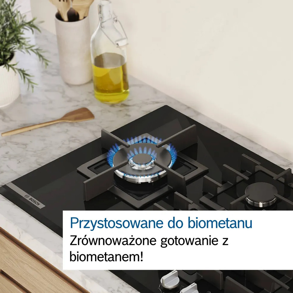 Kuchenka gazowa z zapalonym niebieskim płomieniem. Butelka oleju, drewniana łyżka i rośliny na blacie.