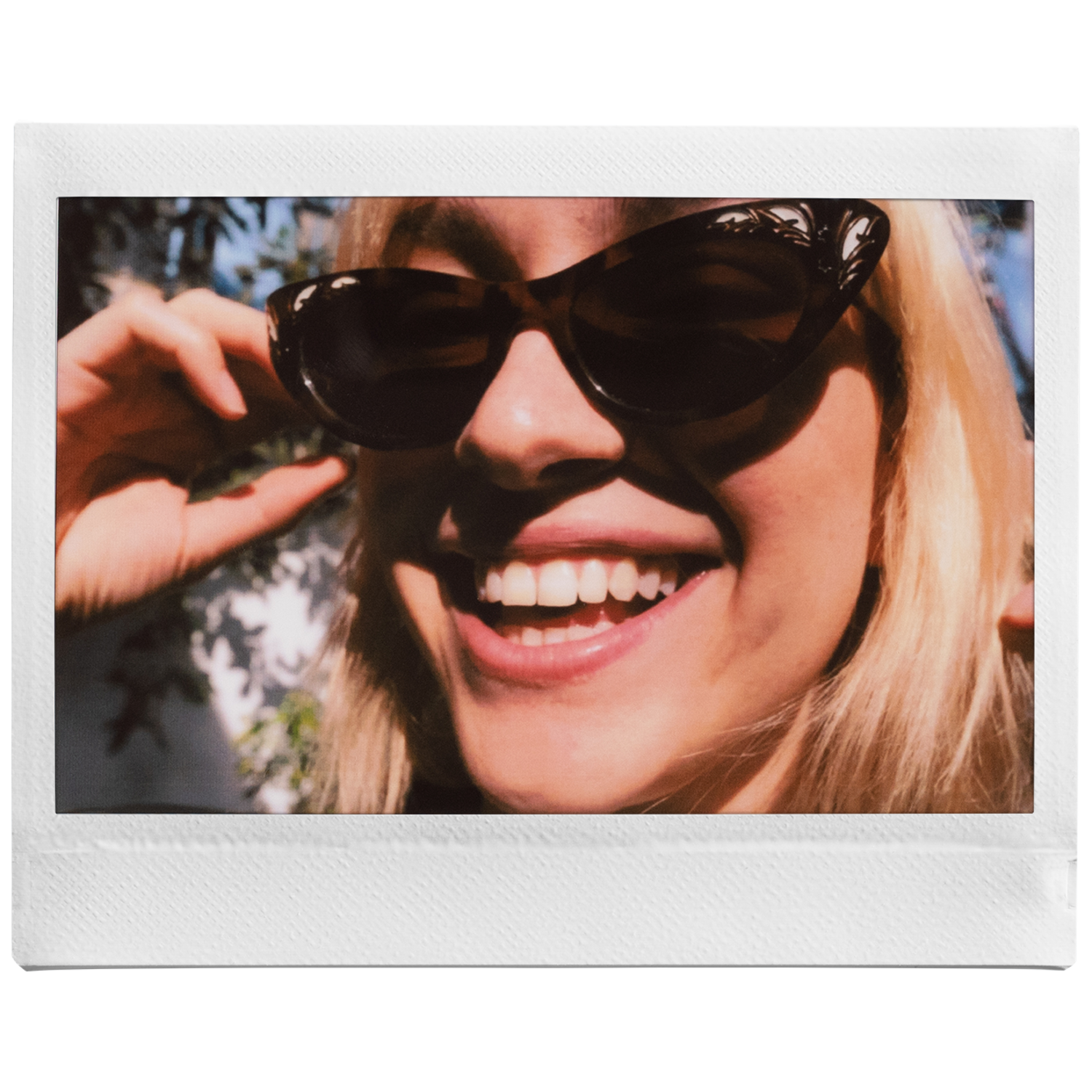 Een vrouw met blond haar en een zonnebril glimlacht breed. De achtergrond heeft wazig groen gebladerte.