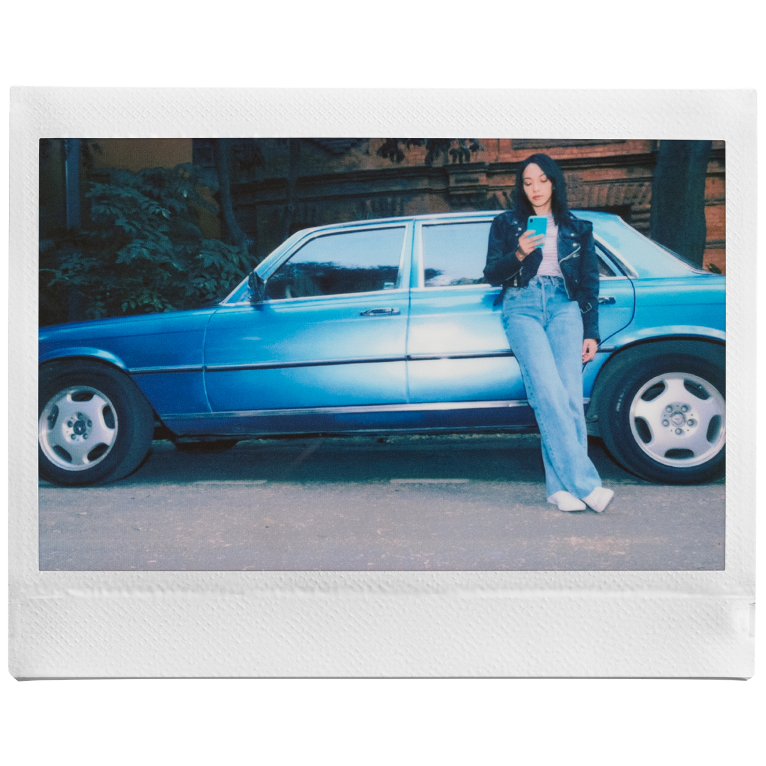 Een vrouw leunt tegen een blauwe auto en kijkt naar haar telefoon. Ze draagt een zwart jasje en een jeans.