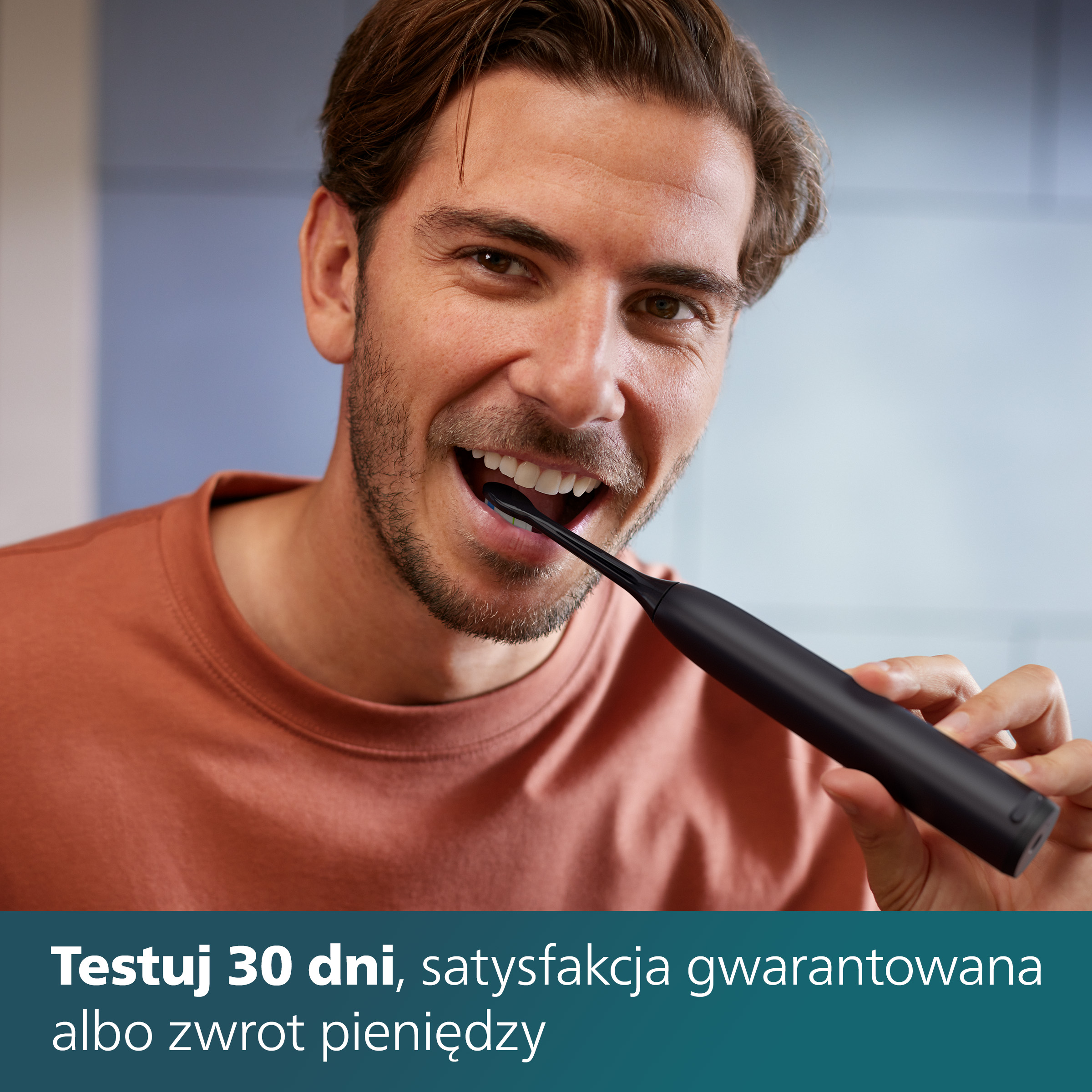 Mężczyzna myjący zęby czarną elektryczną szczoteczką, uśmiechnięty. Pomarańczowa koszula, jasne tło.