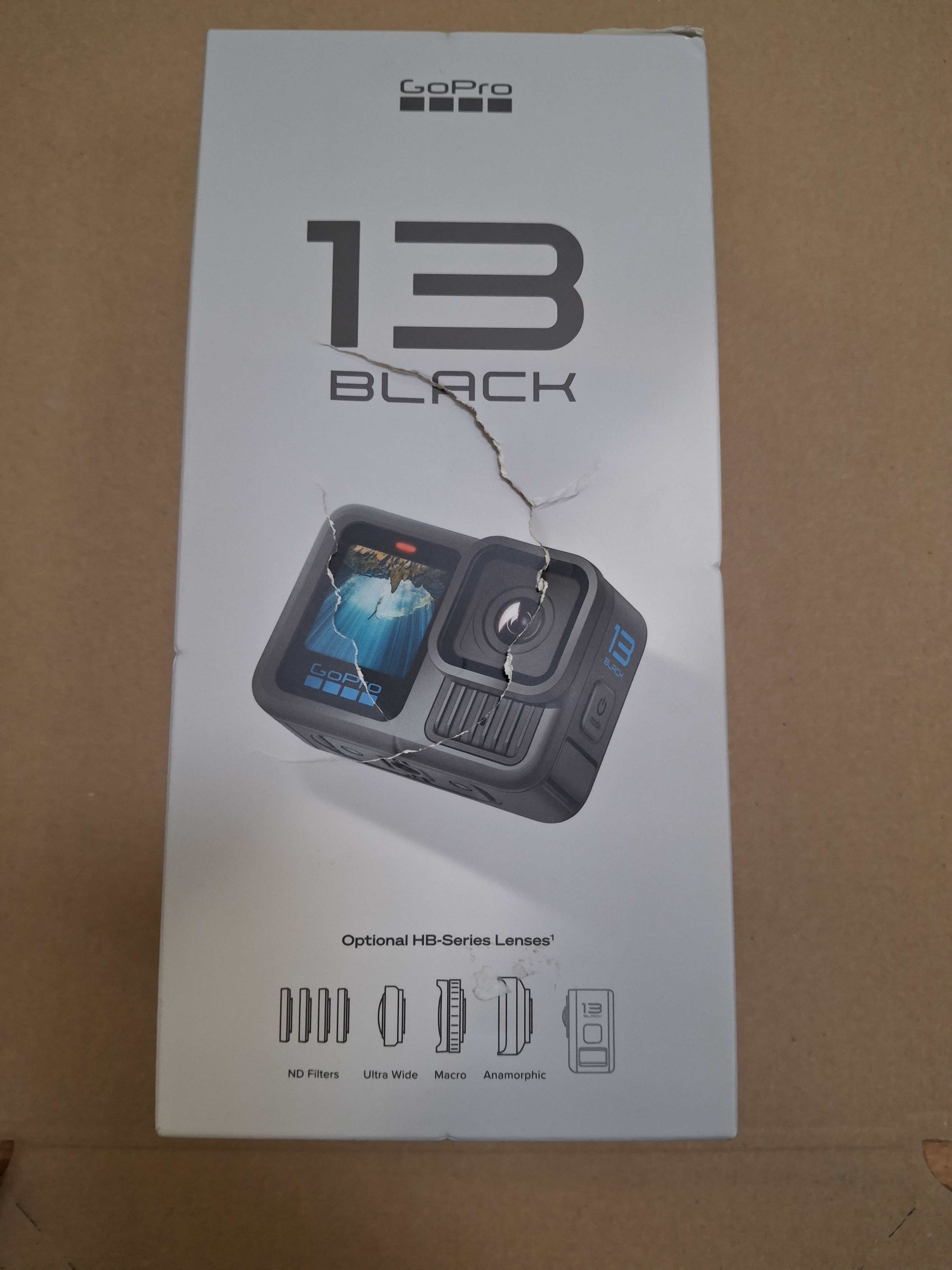 Pudełko GoPro 13 Black ze zdjęciem kamery. Na brązowej powierzchni. Pudełko jest podarte.