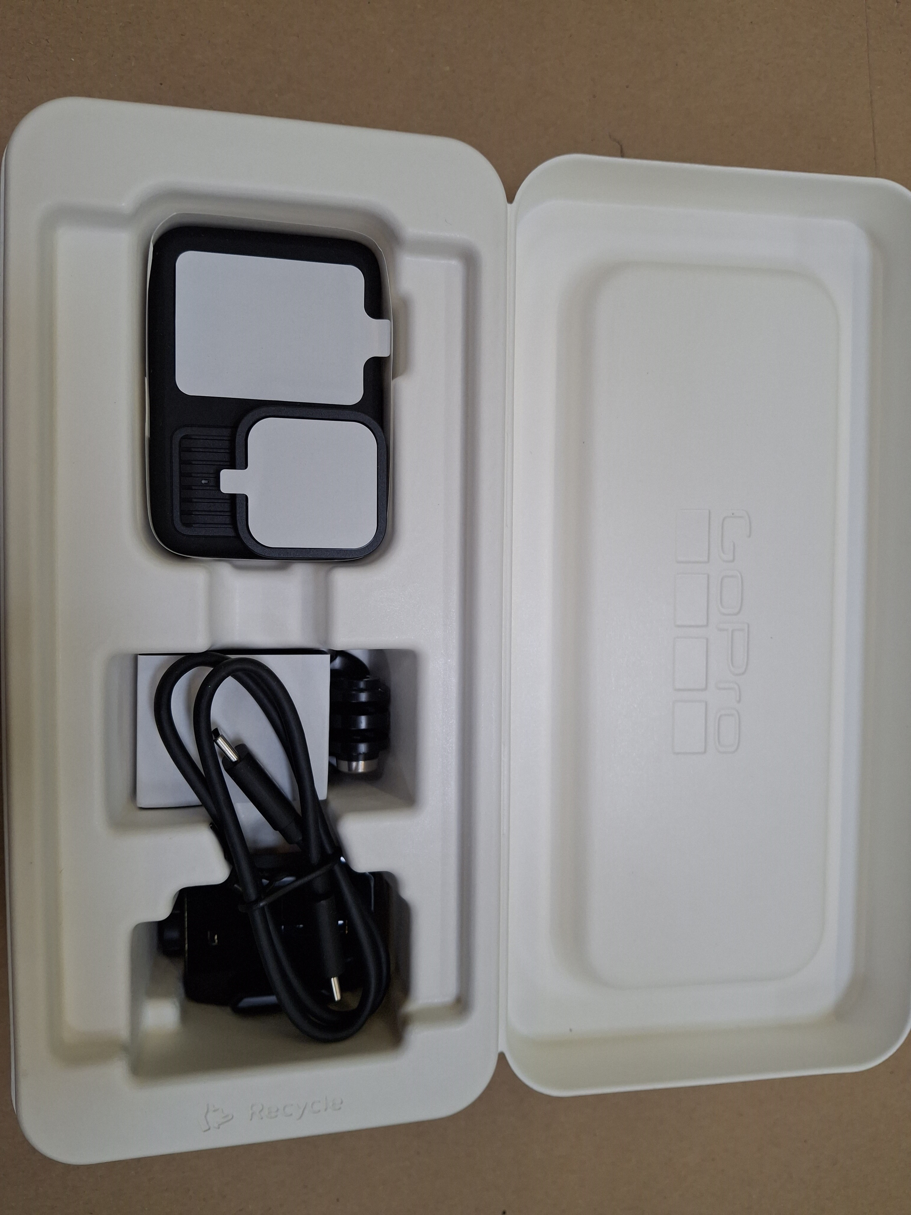 Obudowa GoPro jest otwarta. W środku znajduje się kamera, kabel i akcesoria. Obudowa jest biała, z brązowym tłem.