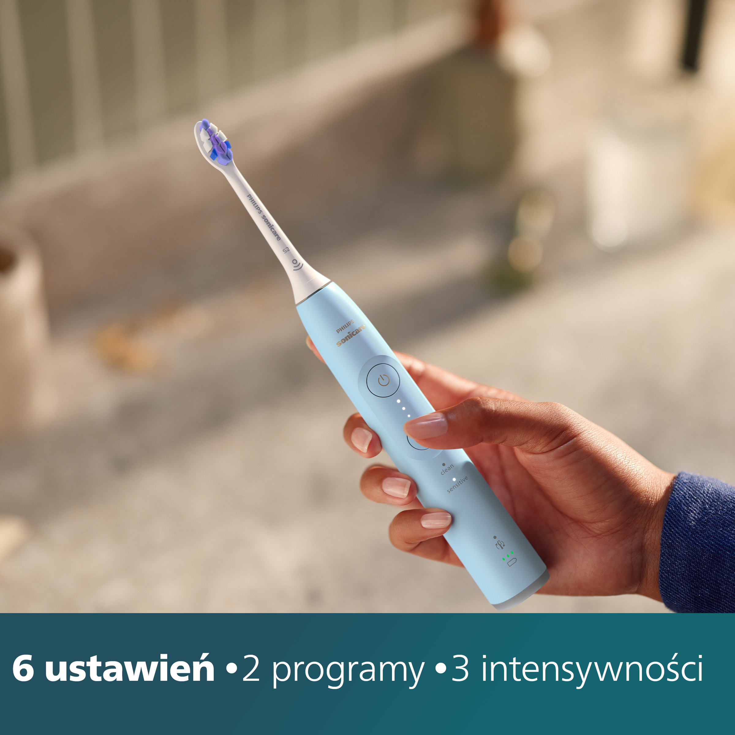 Dłoń trzymająca niebieską szczoteczkę elektryczną. Tekst na dole.