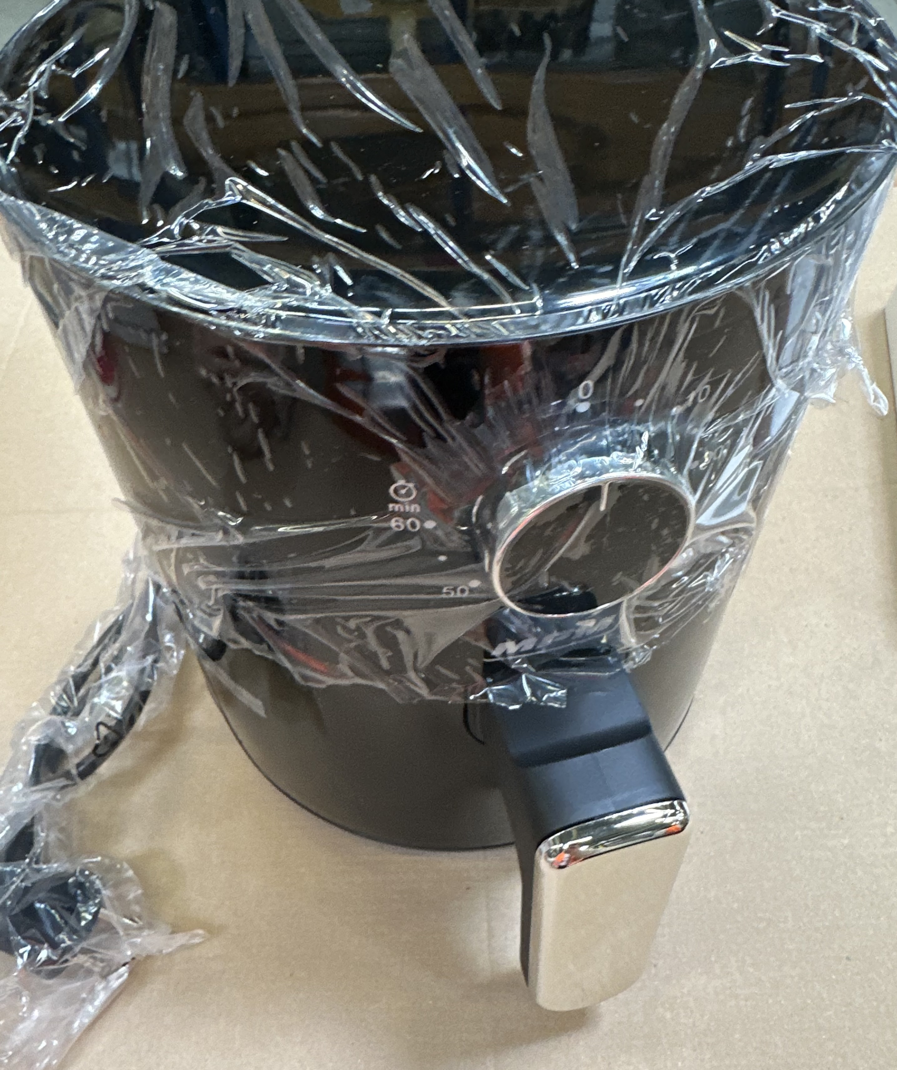 Czarna frytkownica powietrzna w środku. Folia plastikowa częściowo zakrywa frytkownicę. Srebrny uchwyt.