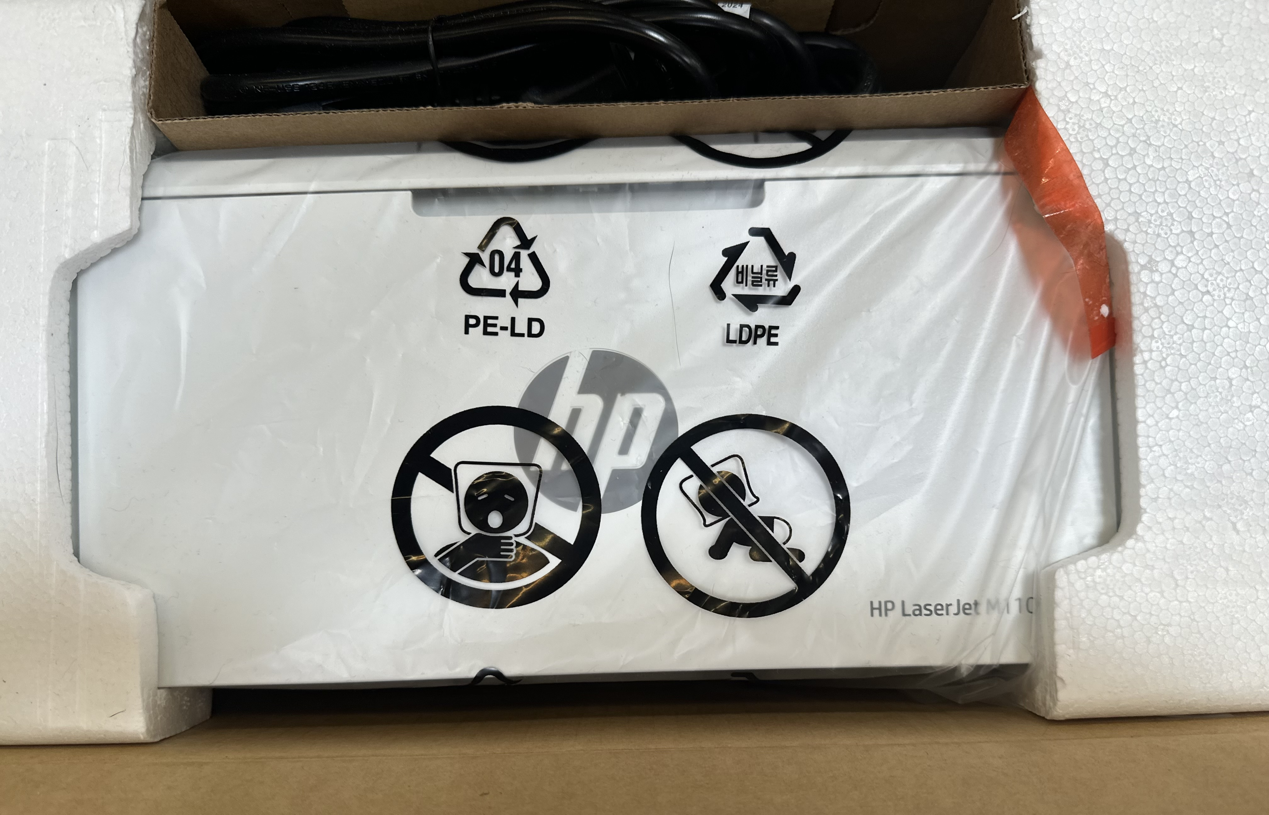 Drukarka HP Laser Jet M110w wewnątrz pudełka. Widoczne symbole bezpieczeństwa i drukarka.