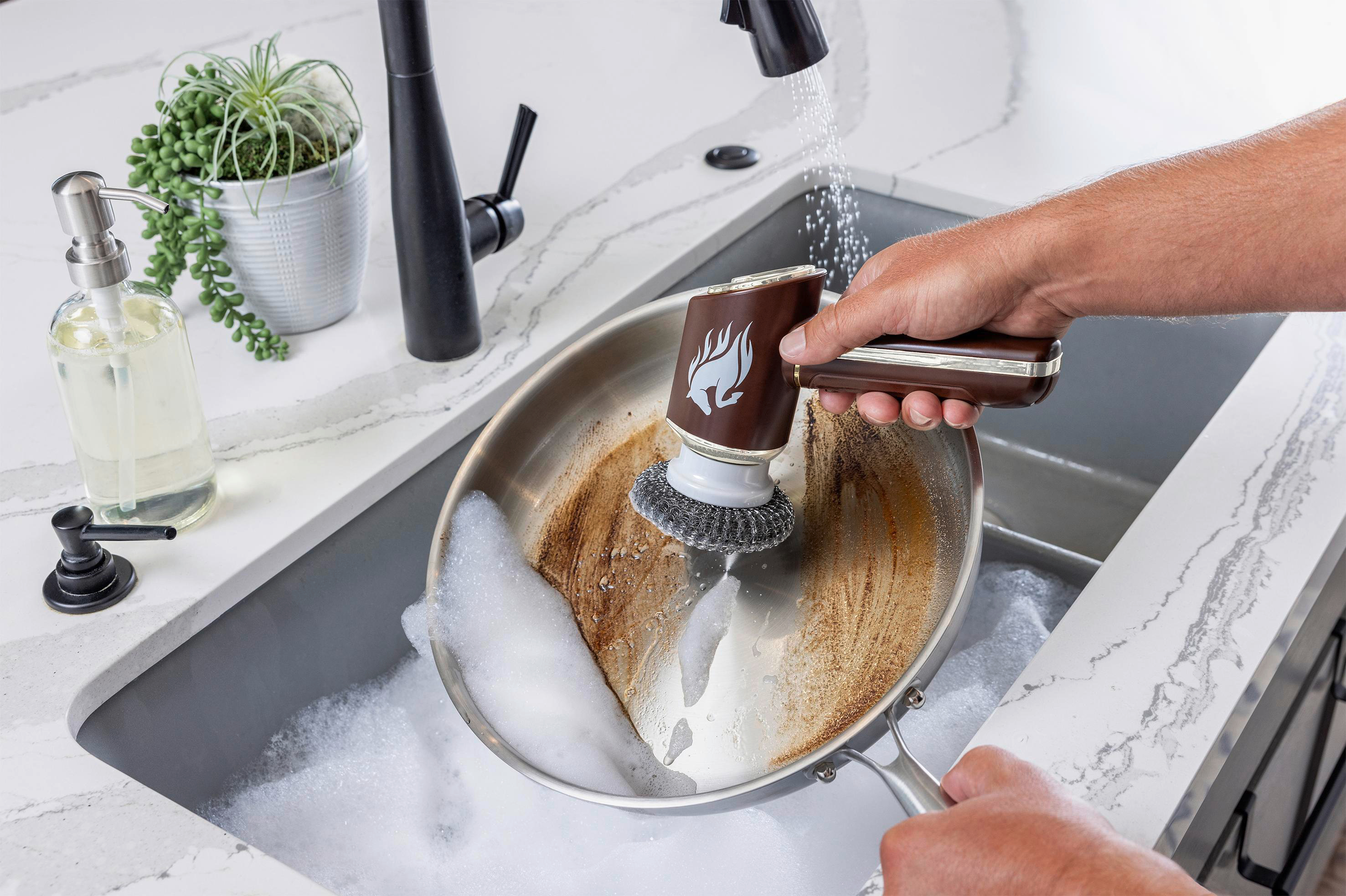 Eine Person reinigt eine schmutzige Edelstahlpfanne mit einem Reinigungswerkzeug in einem Waschbecken mit Wasser.