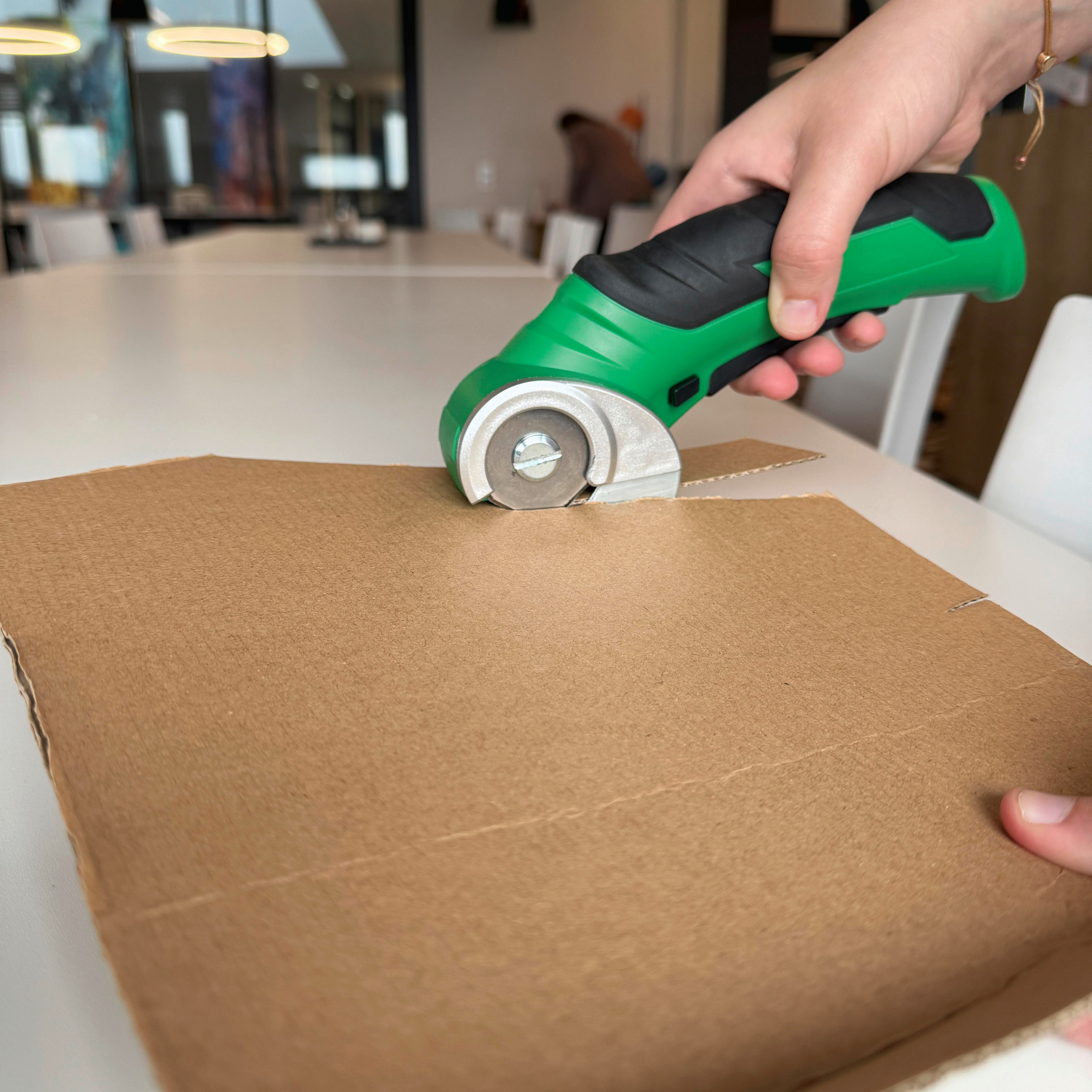 Eine Person, die Pappe mit einem grünen Cutter schneidet. Weißer Tisch, verschwommener Hintergrund.