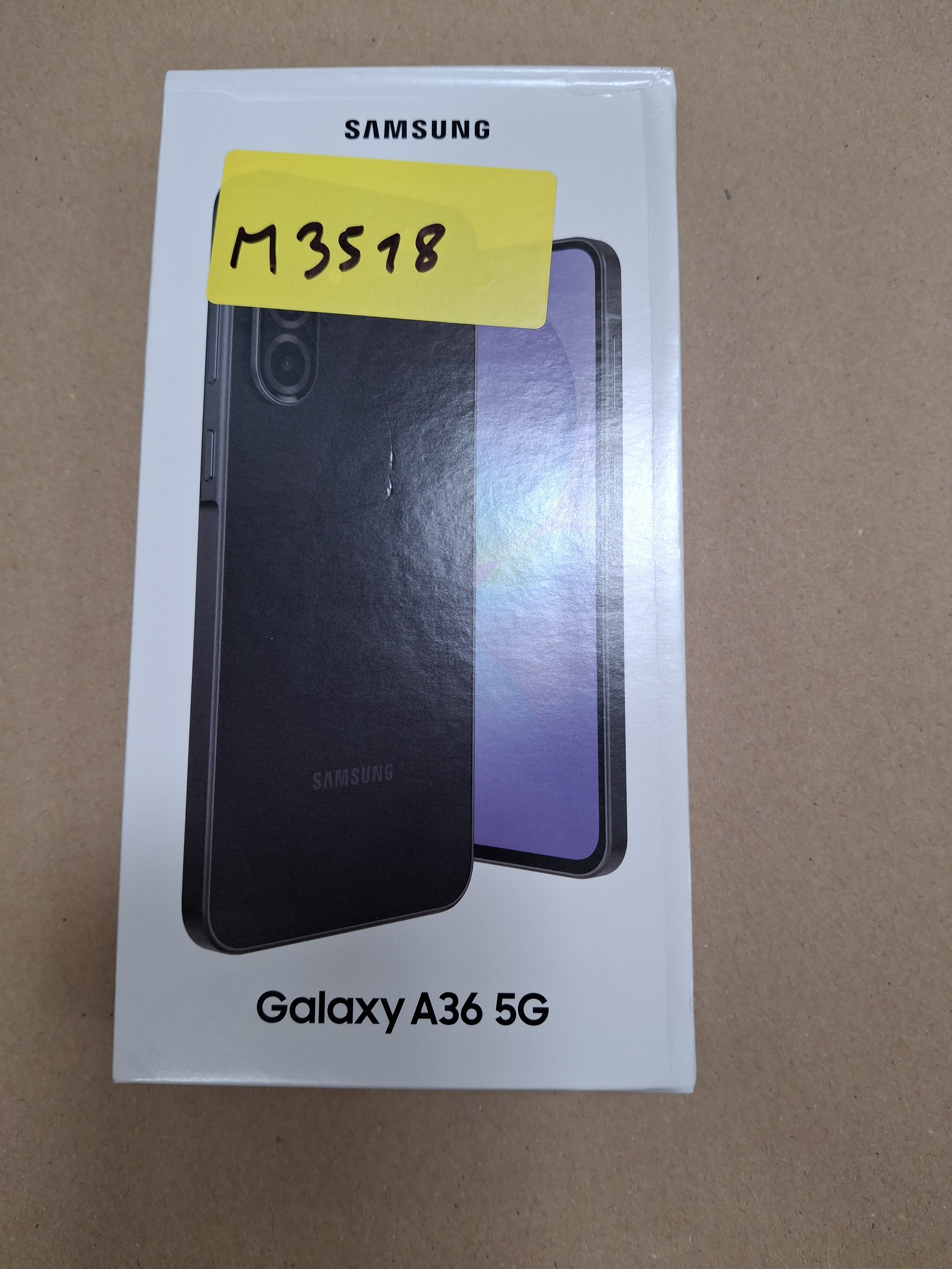 Pudełko telefonu Samsung Galaxy A36 5G. Czarny obraz telefonu, żółta naklejka z "M3518" i logo Samsung. Tło: brązowe.