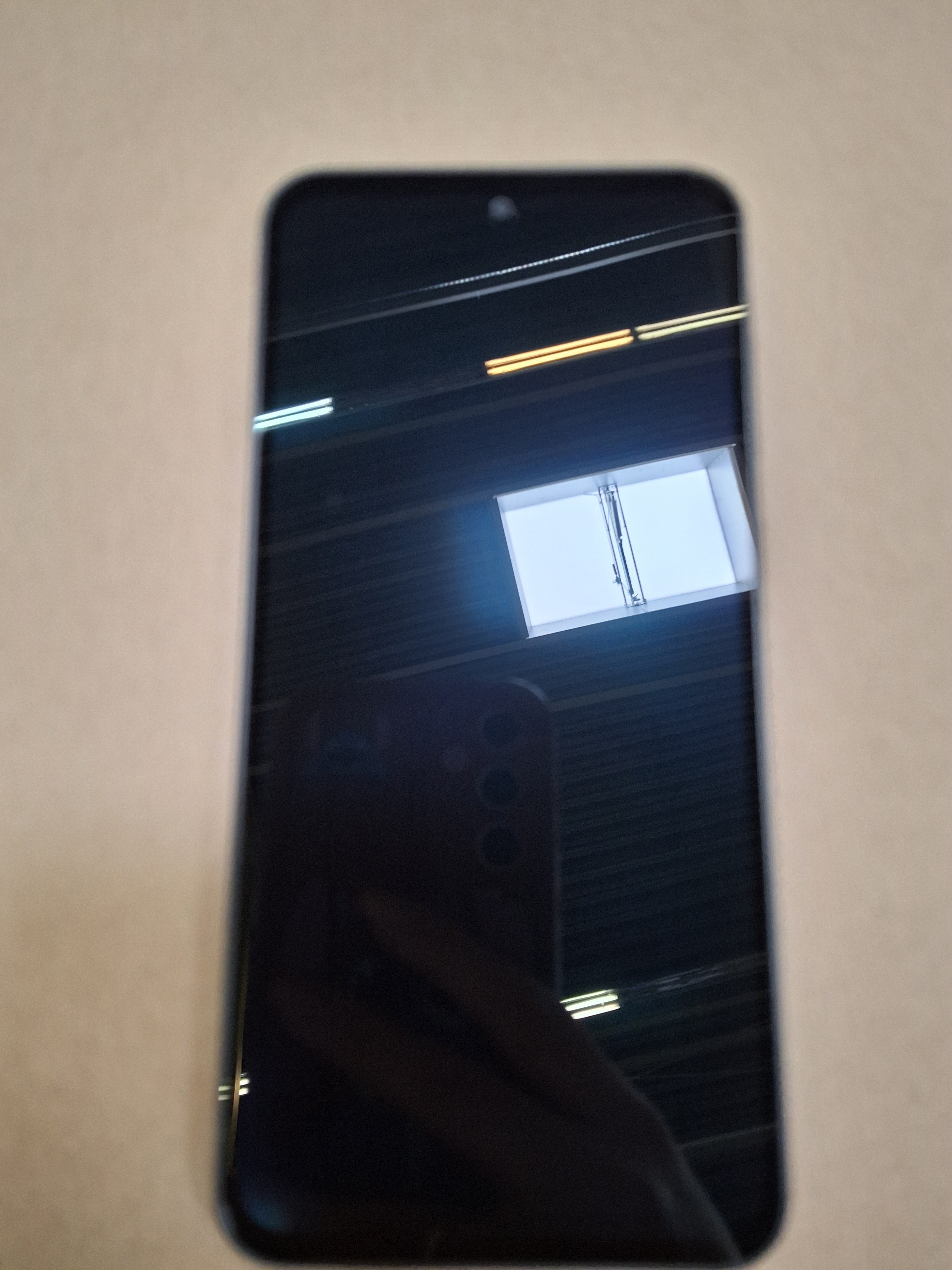 Czarny smartfon z odblaskowym ekranem. Odbicia jasnych świateł sufitowych. Beżowe tło.