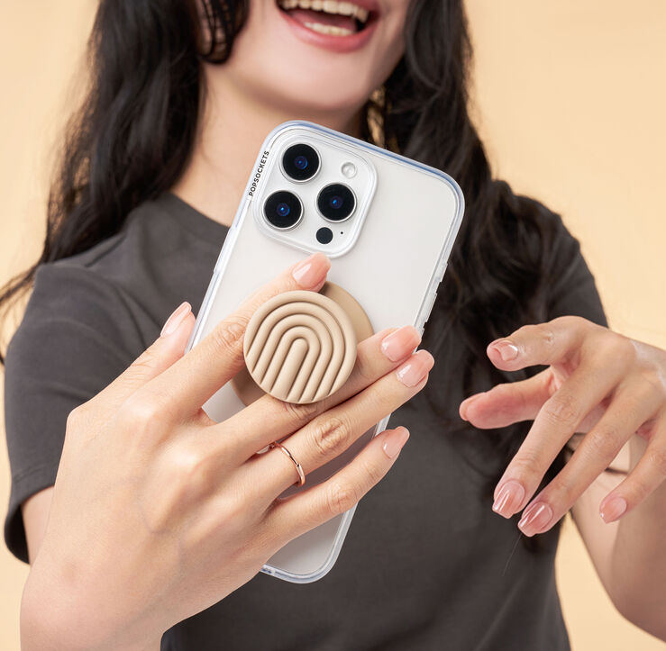 Eine Person hält ein Telefon mit befestigtem hellbraunen PopSockets-Griff, lächelnd, auf beigem Hintergrund.