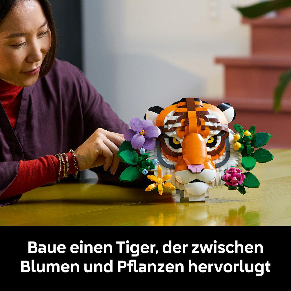 Person baut LEGO-Tigerkopf mit Blumen auf gelber Oberfläche zusammen.