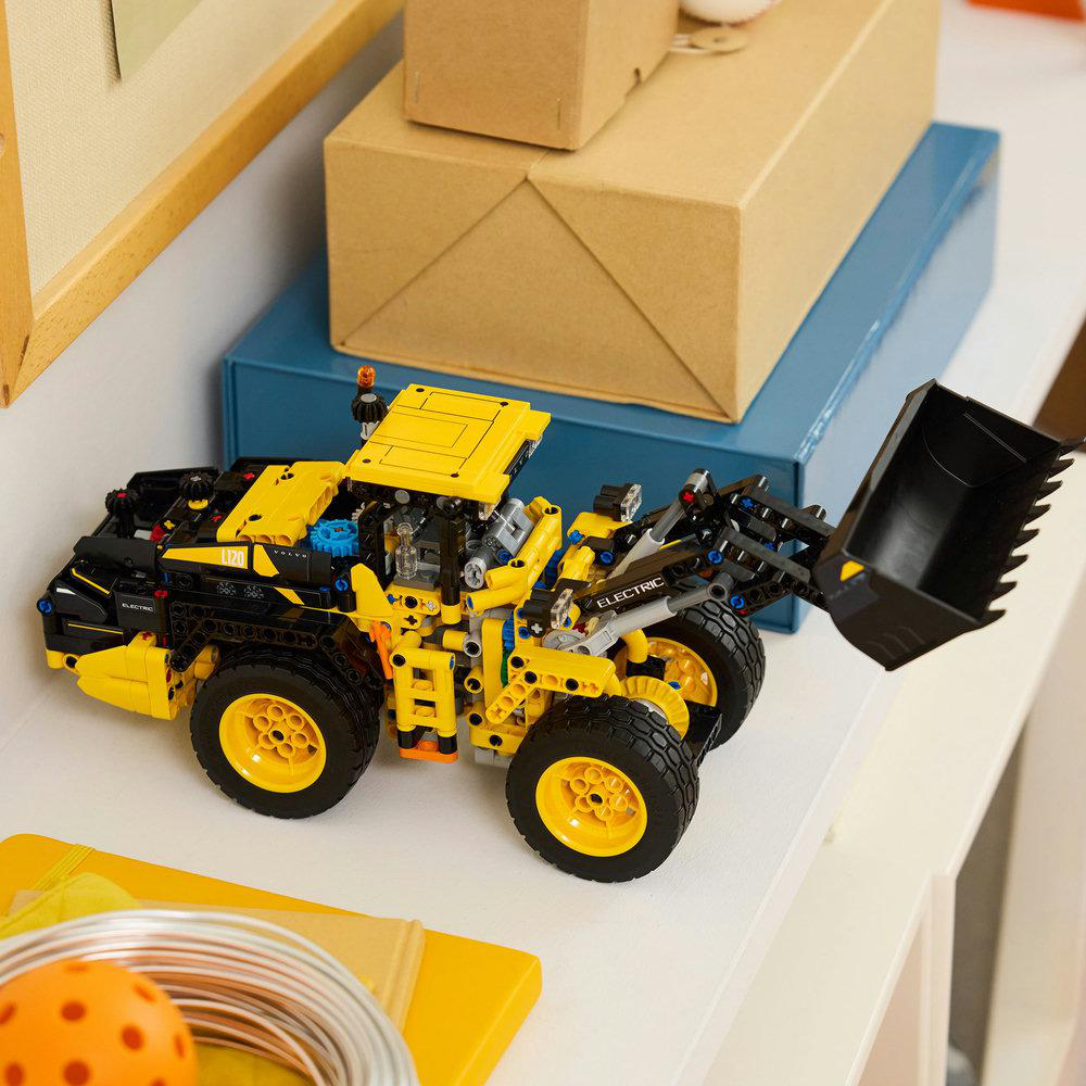 Gelb-schwarzer Lego-Frontlader auf einem weißen Regal, mit Kisten im Hintergrund.