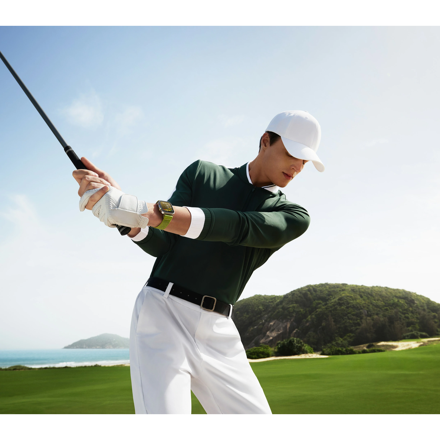 Man in golf attire, swinging club. Wearing white cap, shirt, and pants. Green watch on wrist.