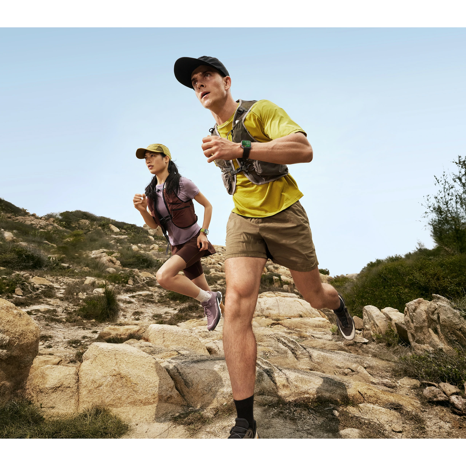 Two runners on a rocky trail. Man and woman running uphill. Both wearing running gear and smartwatches.