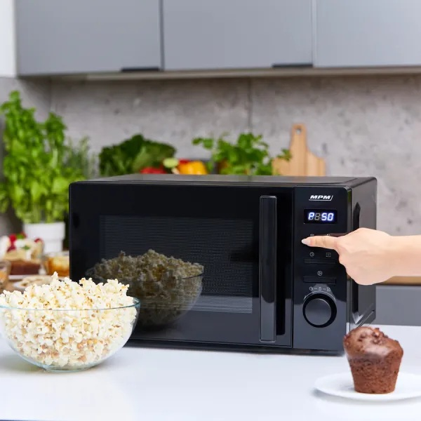 Czarna kuchenka mikrofalowa z popcornem i babeczką. Ręka naciska przycisk.