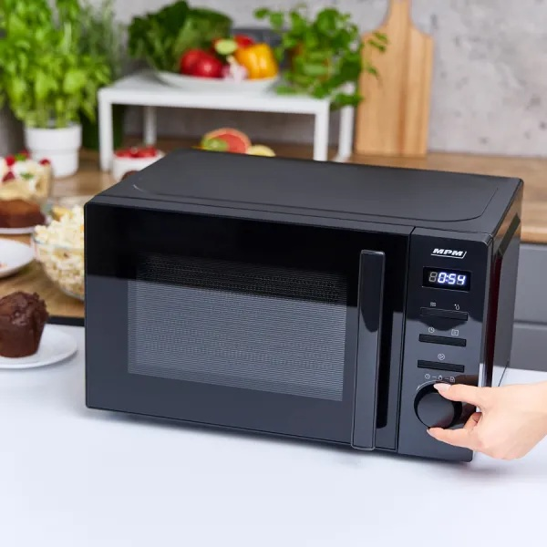 Czarna kuchenka mikrofalowa. Ręka przekręca pokrętło. Jedzenie i rośliny w tle.