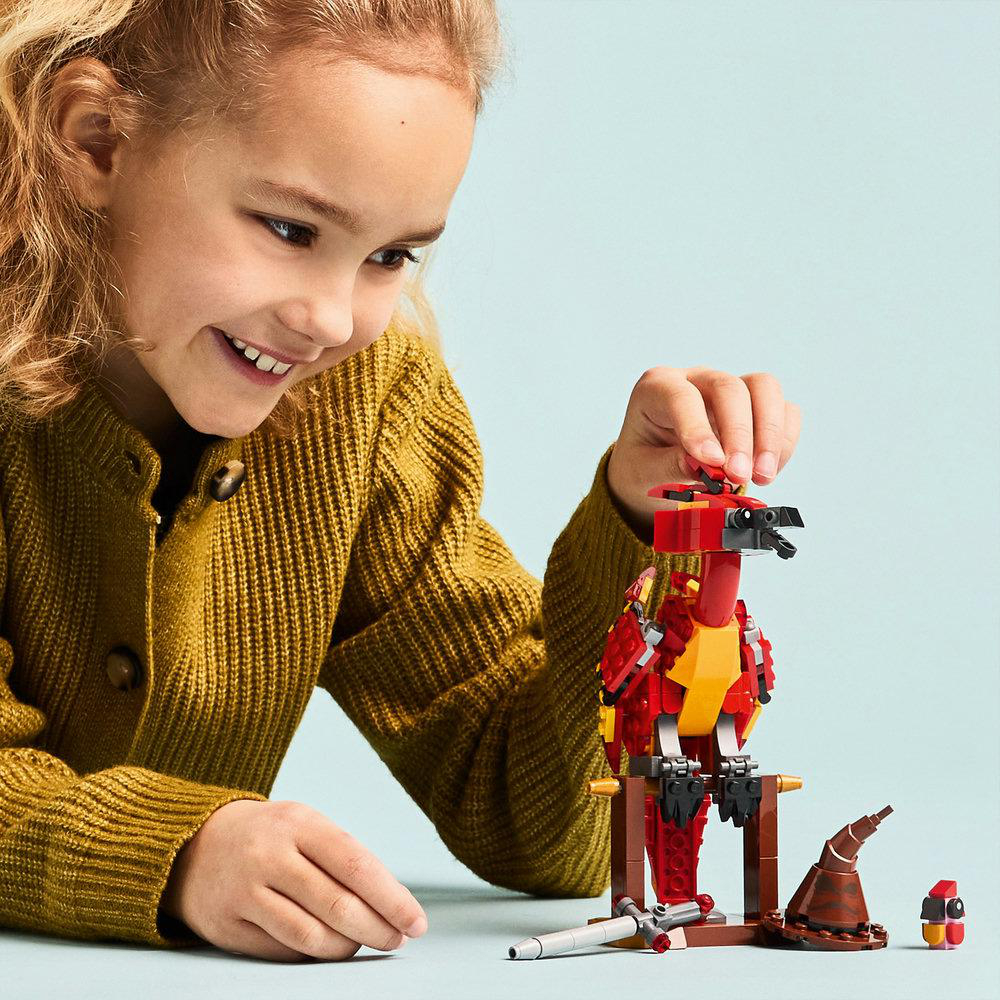 Ein Mädchen spielt mit einem roten und gelben Lego-Vogelmodell. Ein brauner Hut und ein Zauberstab sind in der Nähe.