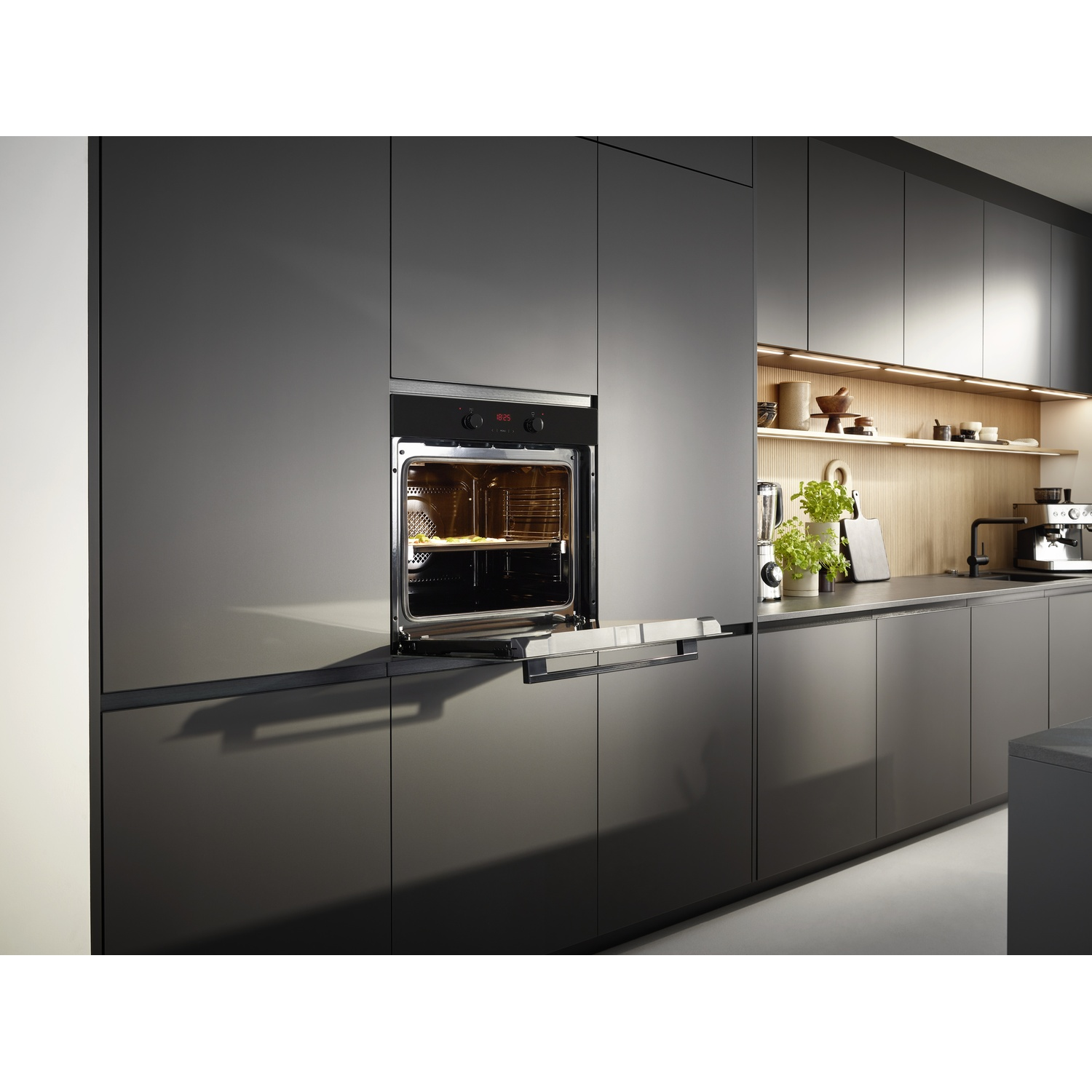 Cucina moderna con forno aperto, mostra cibo. Mobili grigio scuro, mensola in legno con piante.