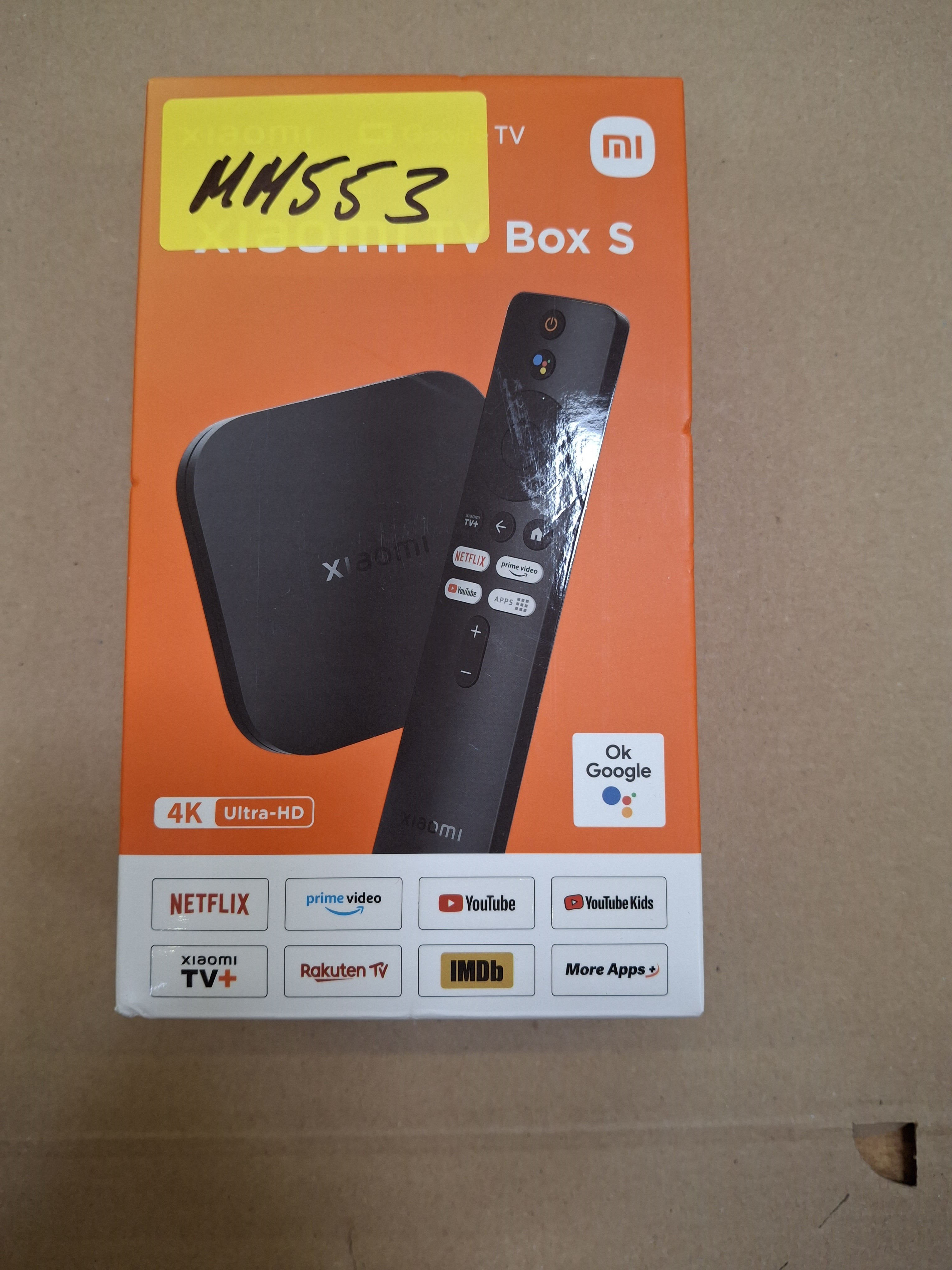 Pomarańczowe pudełko z Xiaomi TV Box i pilotem. Zawiera logo serwisów streamingowych i żółtą etykietę.