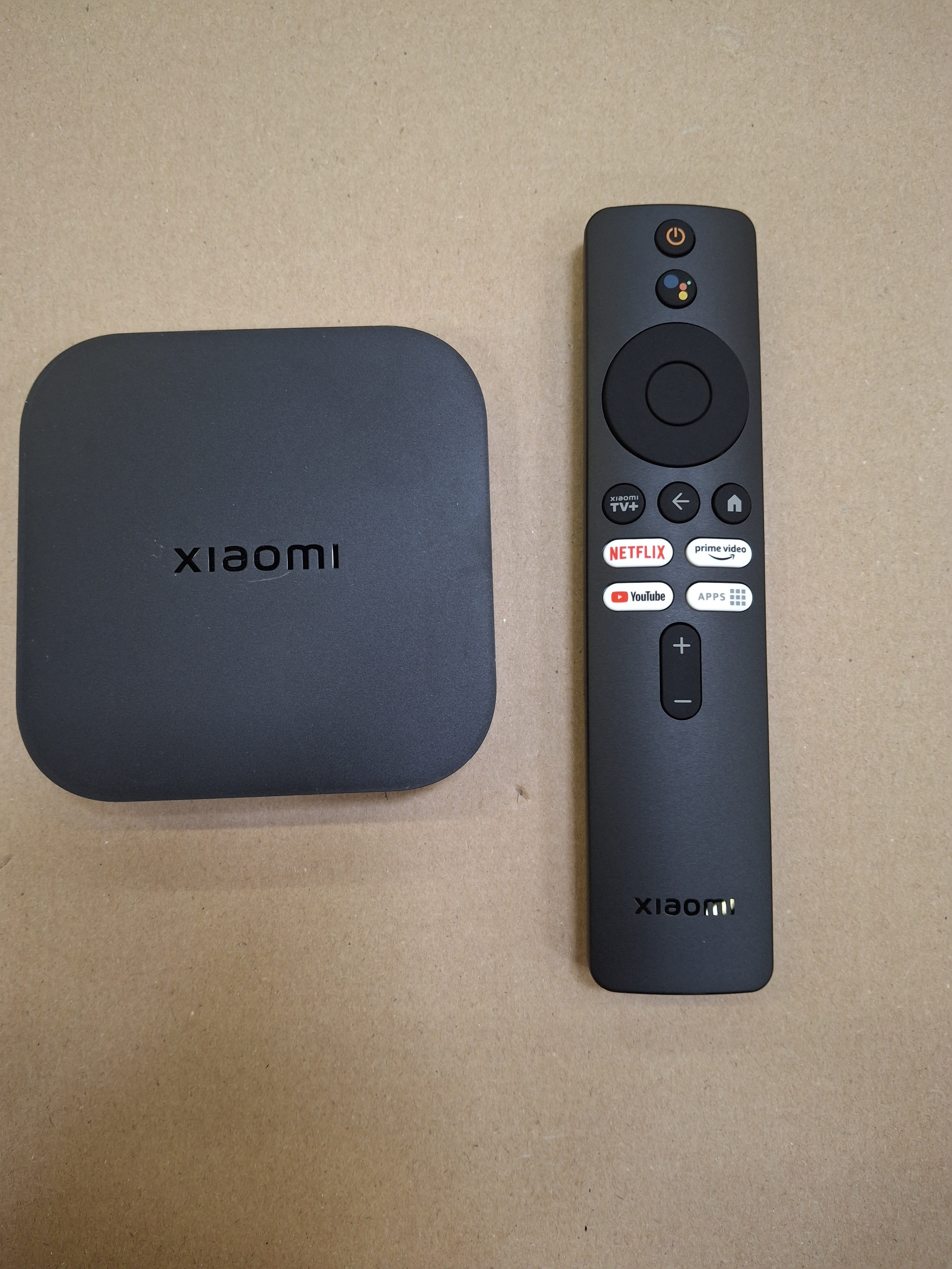 Xiaomi TV Box i pilot na beżowej powierzchni. Pudełko jest kwadratowe, a pilot długi i czarny.