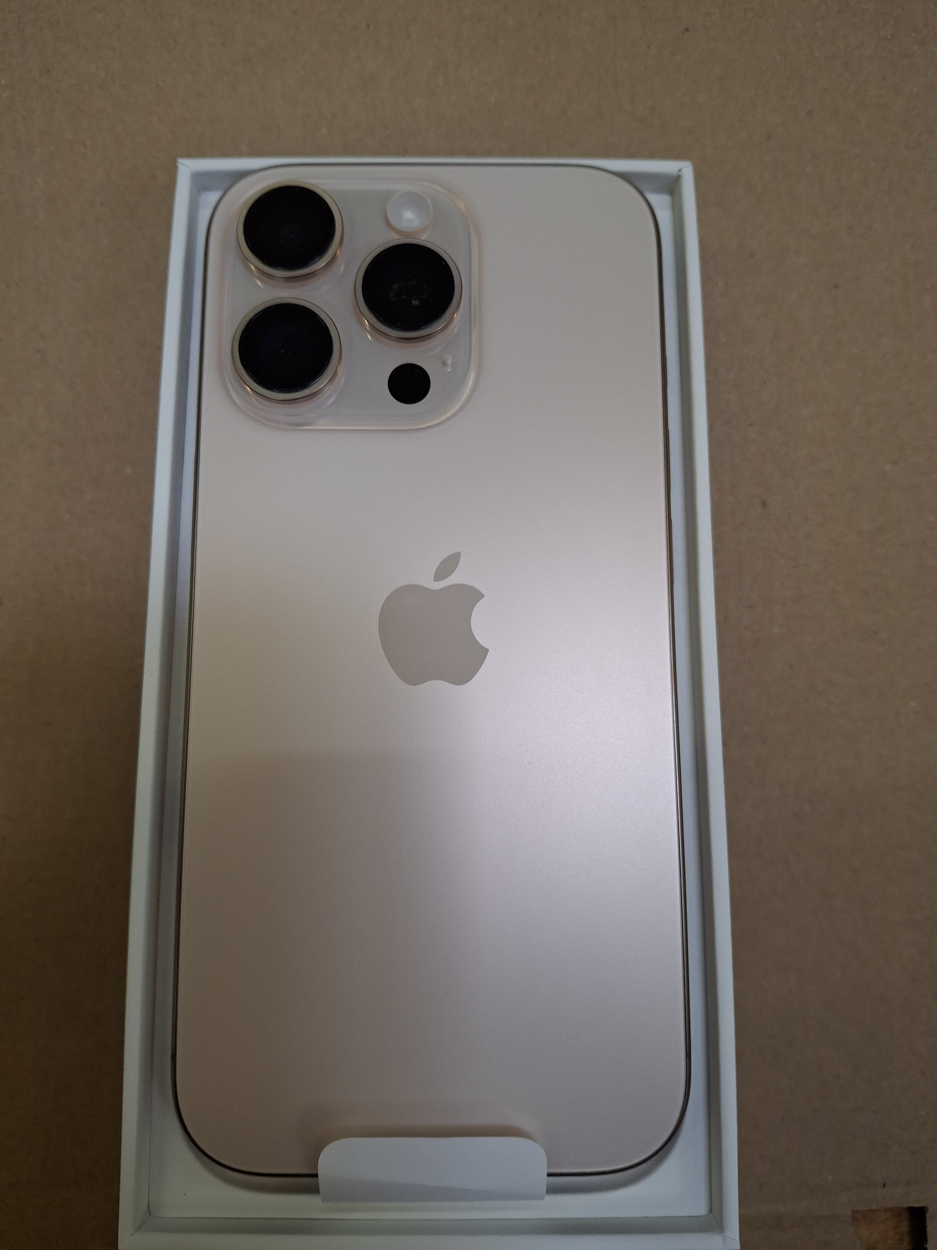 Nowy złoty iPhone w białym pudełku. Telefon ma potrójny obiektyw i logo Apple.