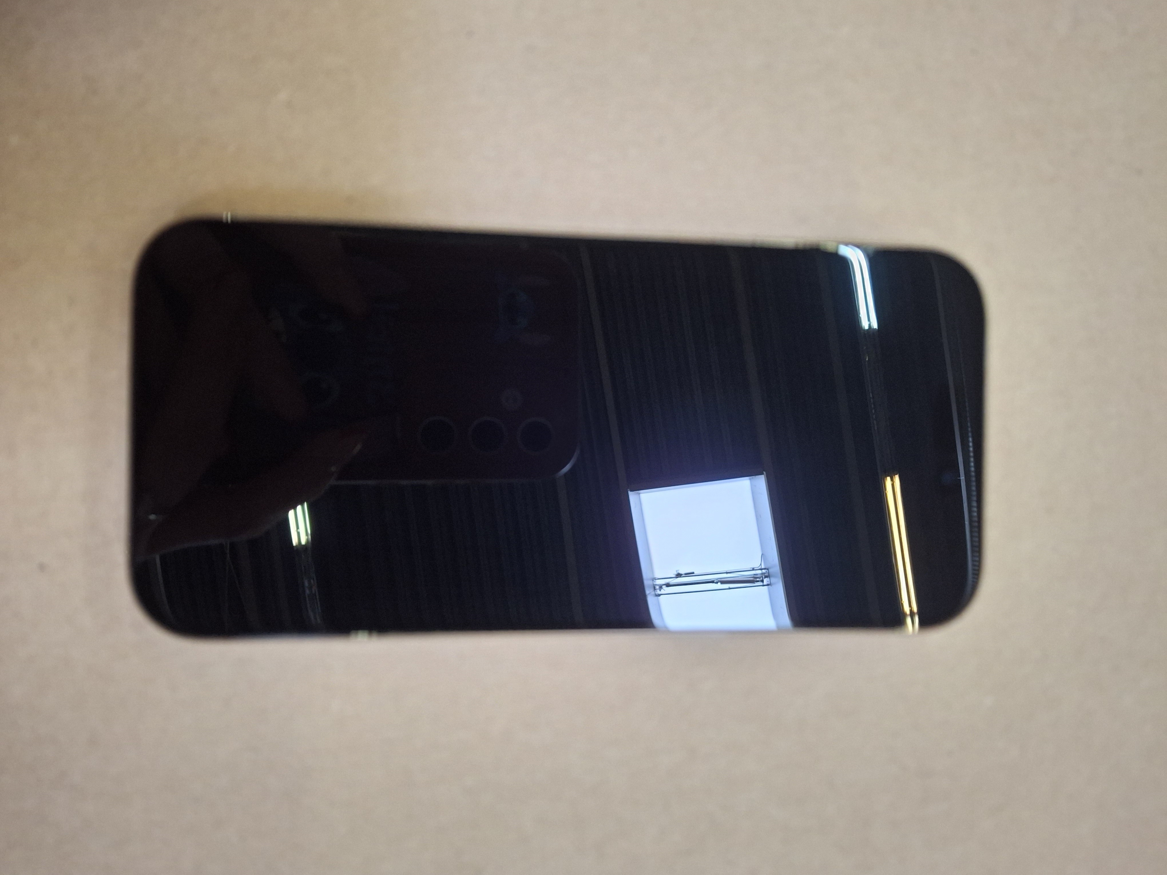 Czarny telefon odbijający okno i inne obiekty na jasnobrązowej powierzchni. Telefon ma potrójny aparat.