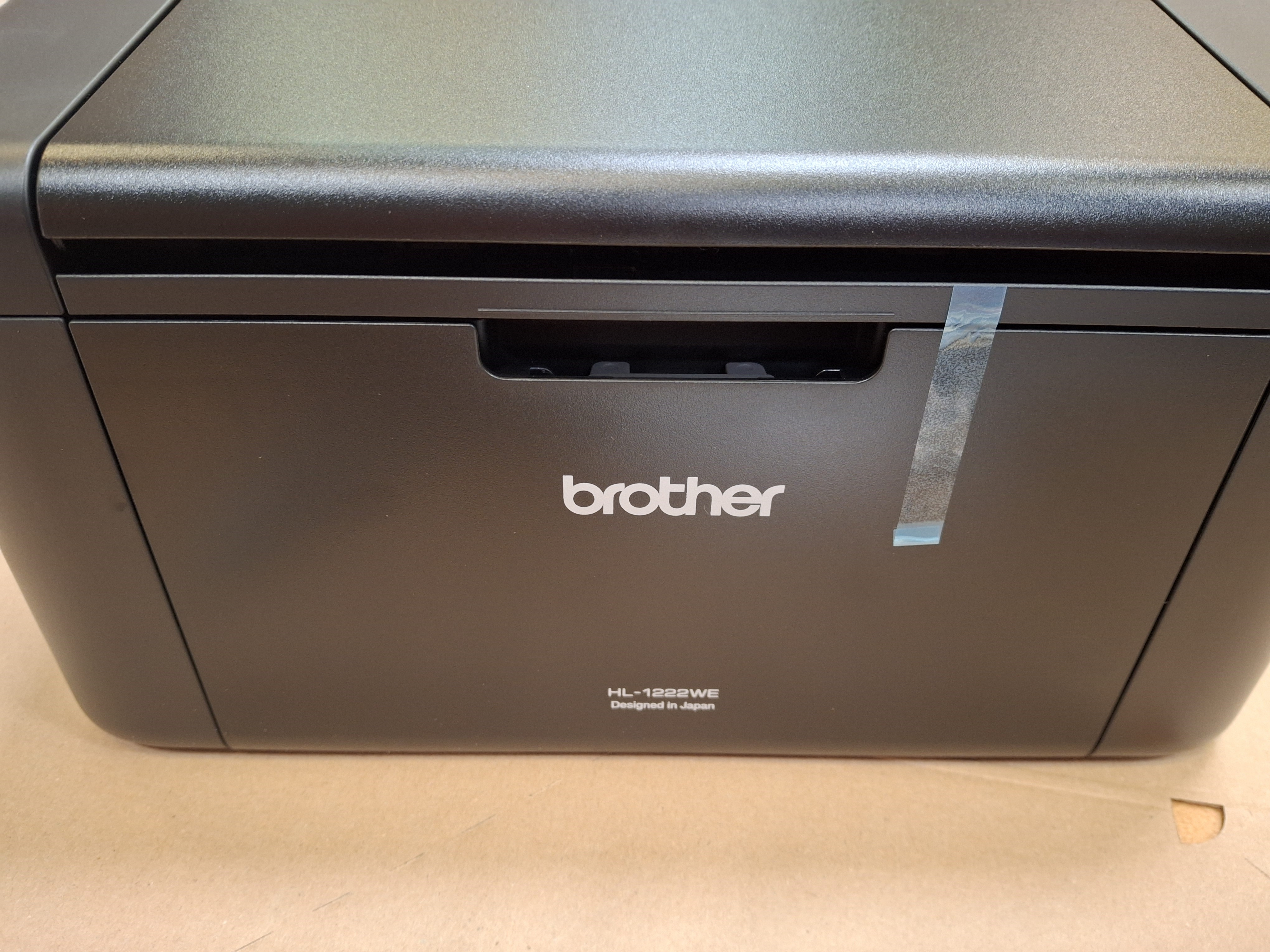 Czarna drukarka Brother. Przód ma logo, numer modelu i taśmę. Na jasnobrązowej powierzchni.