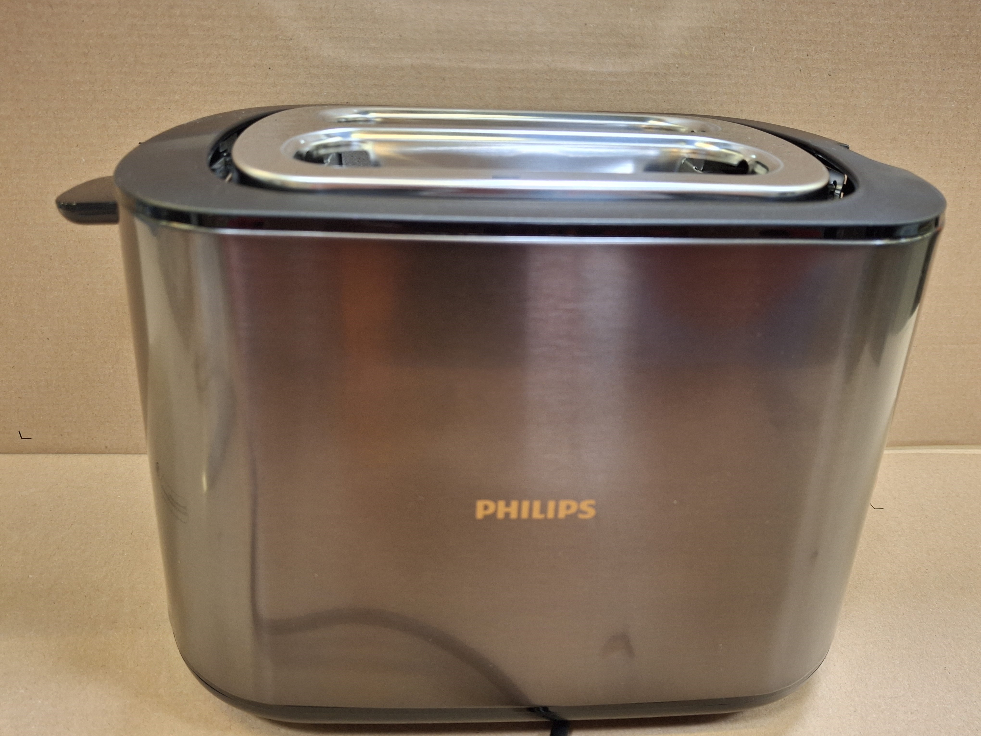 Metalowy toster Philips jest wyśrodkowany. Ma czarny wierzch, srebrną szczelinę i stoi na brązowym kartonie.