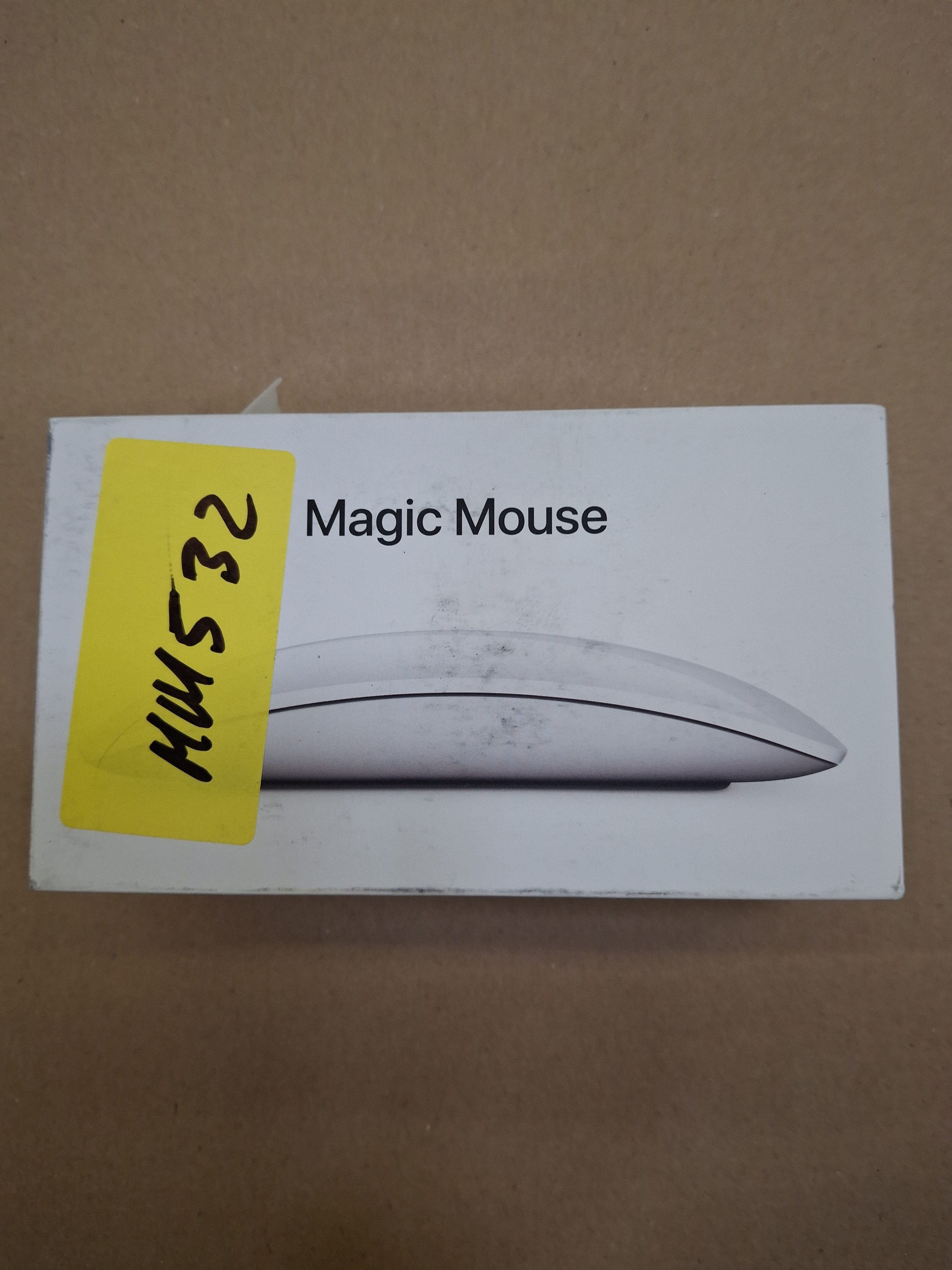 Zapakowana mysz Apple Magic Mouse z żółtą naklejką na brązowej powierzchni. Etykieta: Magic Mouse.