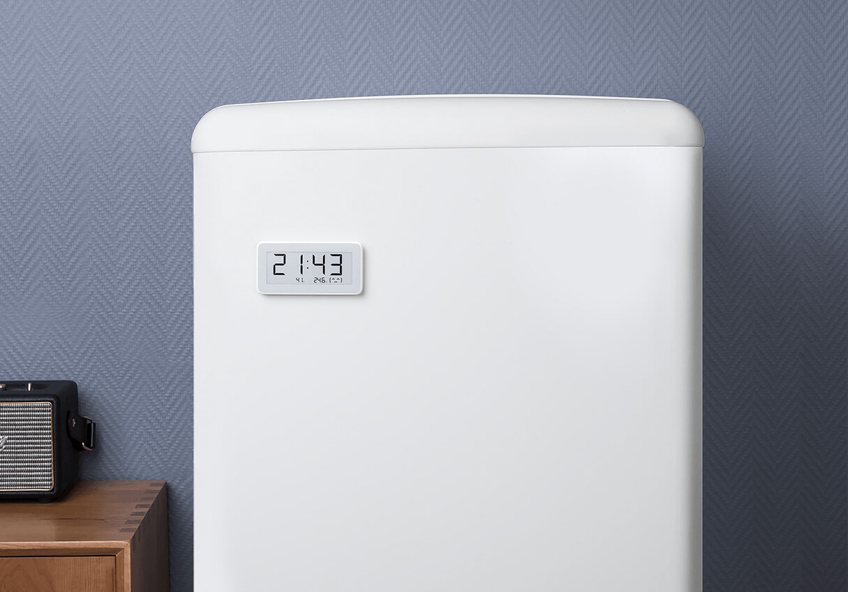 Białe urządzenie z cyfrowym zegarem pokazującym 21:43. Szara ściana w tle, drewniany stół i czarny głośnik po lewej.