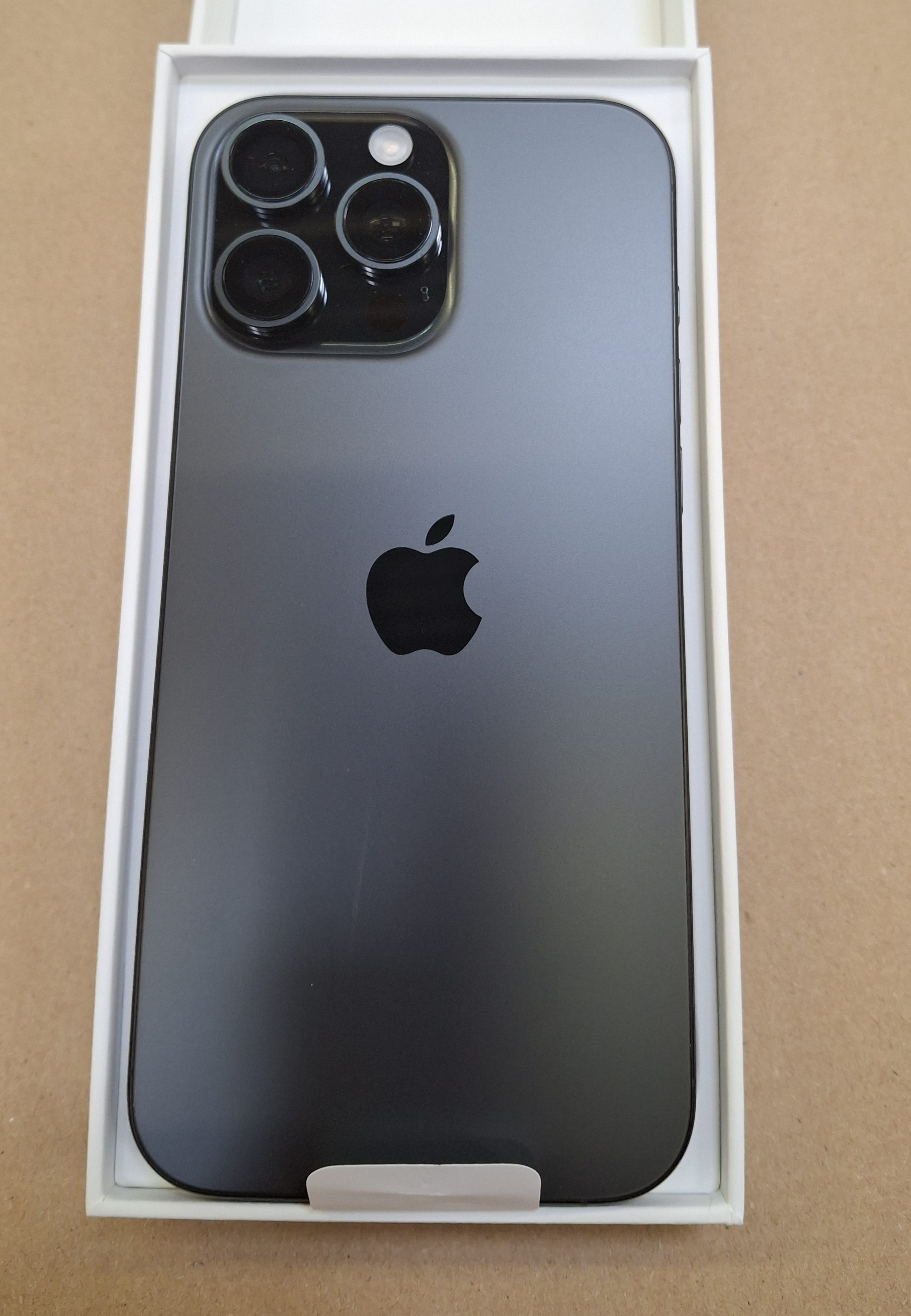 Czarny iPhone w białym pudełku. Trzy kamery, logo Apple. Tło: jasnobrązowa powierzchnia.